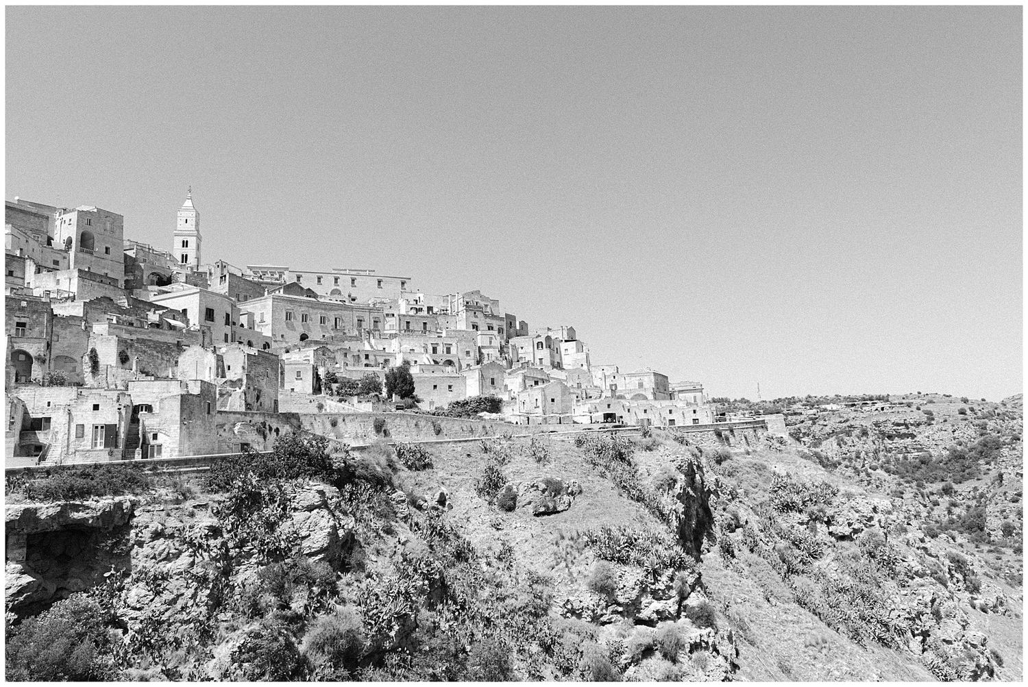 Black and white photograph showing the ancient hillside city of Matera, Italy with stone buildings cascading down the rocky slope.