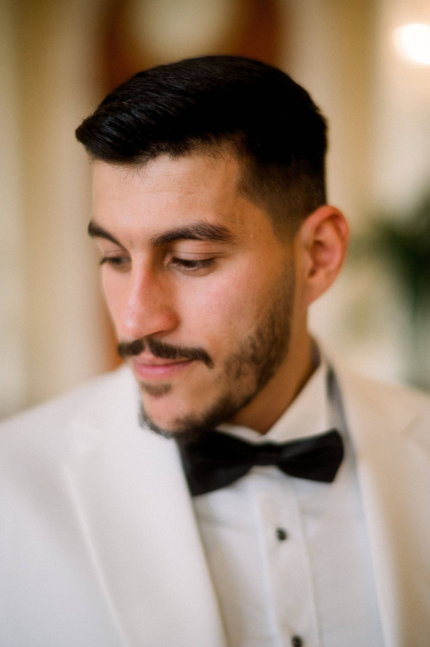 Close-up portrait of a groom in a white tuxedo jacket and black bow tie looking down with a thoughtful expression.
