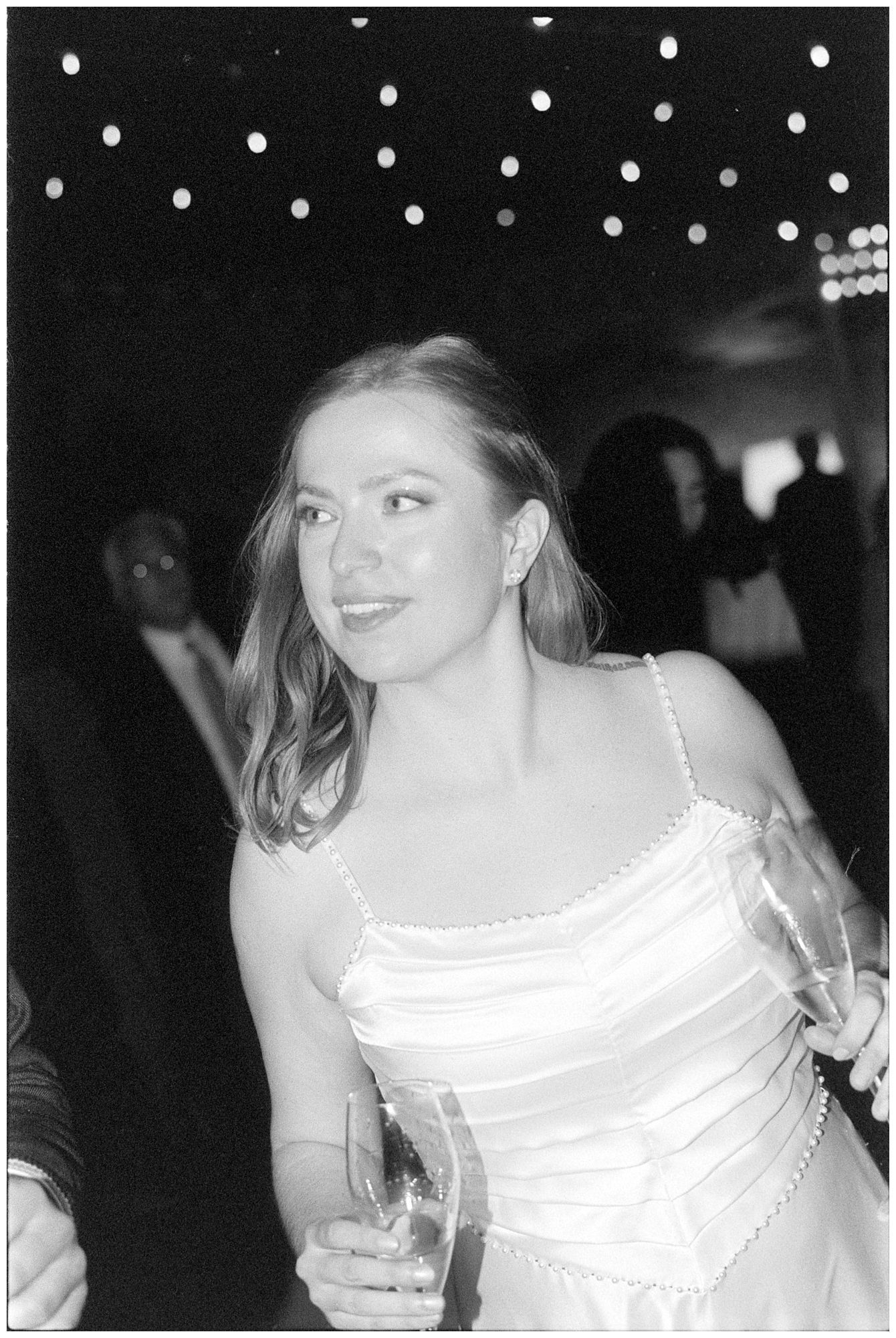 A black and white photo of someone in a white dress at a party with string lights overhead.