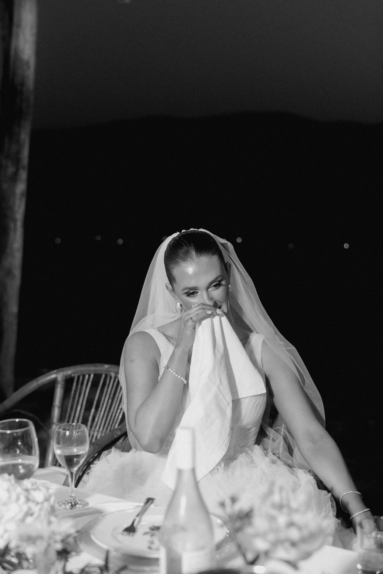 A bride in a white wedding dress and veil sits at a dinner table with glassware and flowers in black and white photography.
