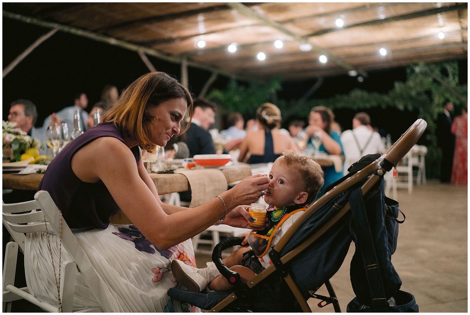 A parent gently feeding their child indoors at an evening event with dining tables and ambient lighting.