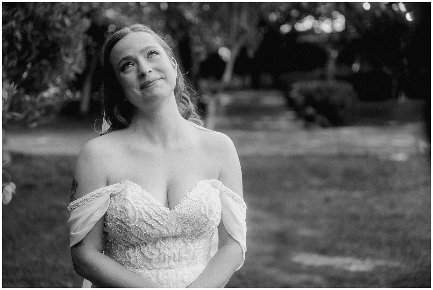 Black and white portrait of a bride in an off-shoulder lace wedding dress smiling with a joyful expression outdoors.