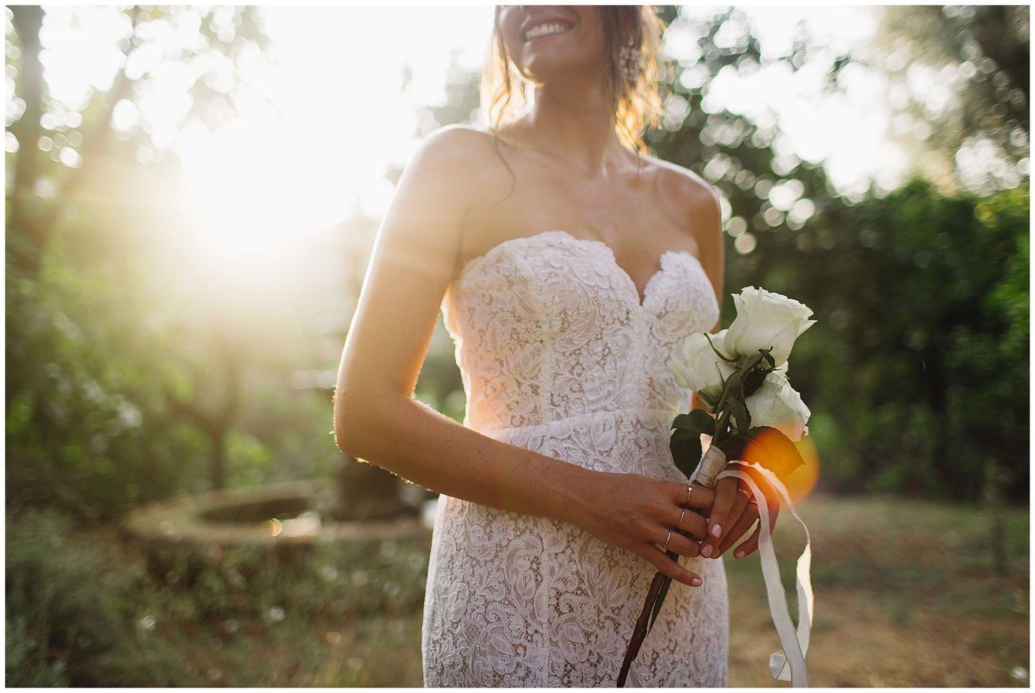 A bride in a lace strapless wedding dress holds a white bouquet during golden hour sunset in a garden setting.