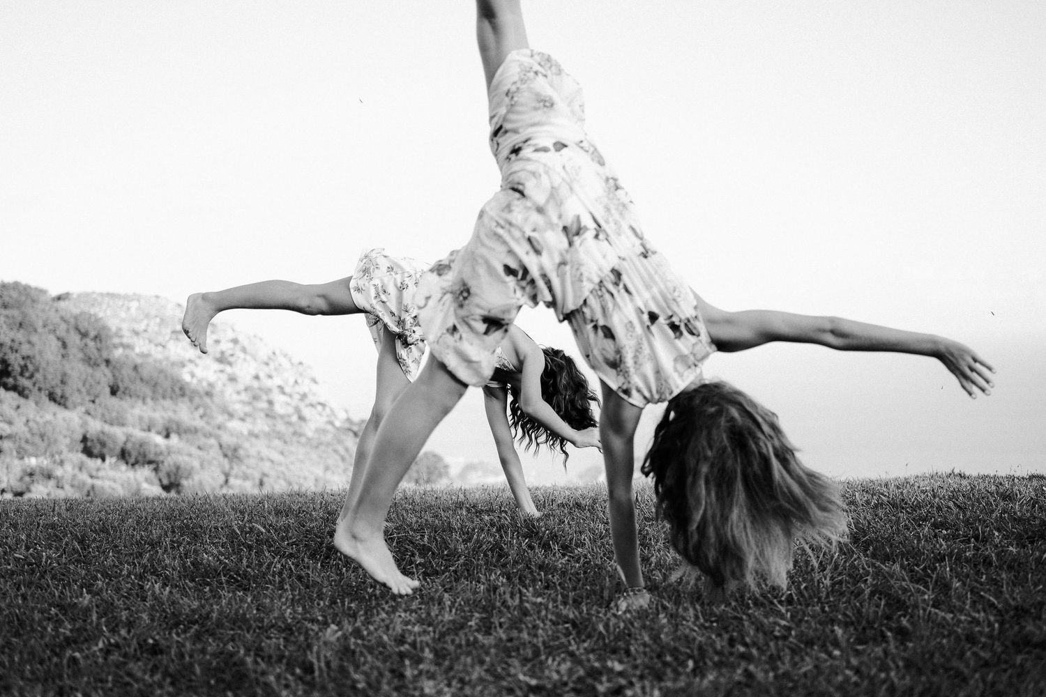 Black and white photo of person doing a cartwheel on grassy hill wearing floral dress in summer.