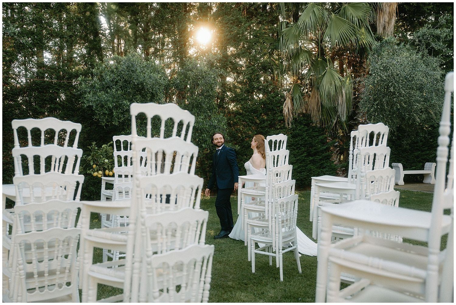White wooden chairs arranged for an outdoor event in a forest setting with sunlight peeking through the trees.