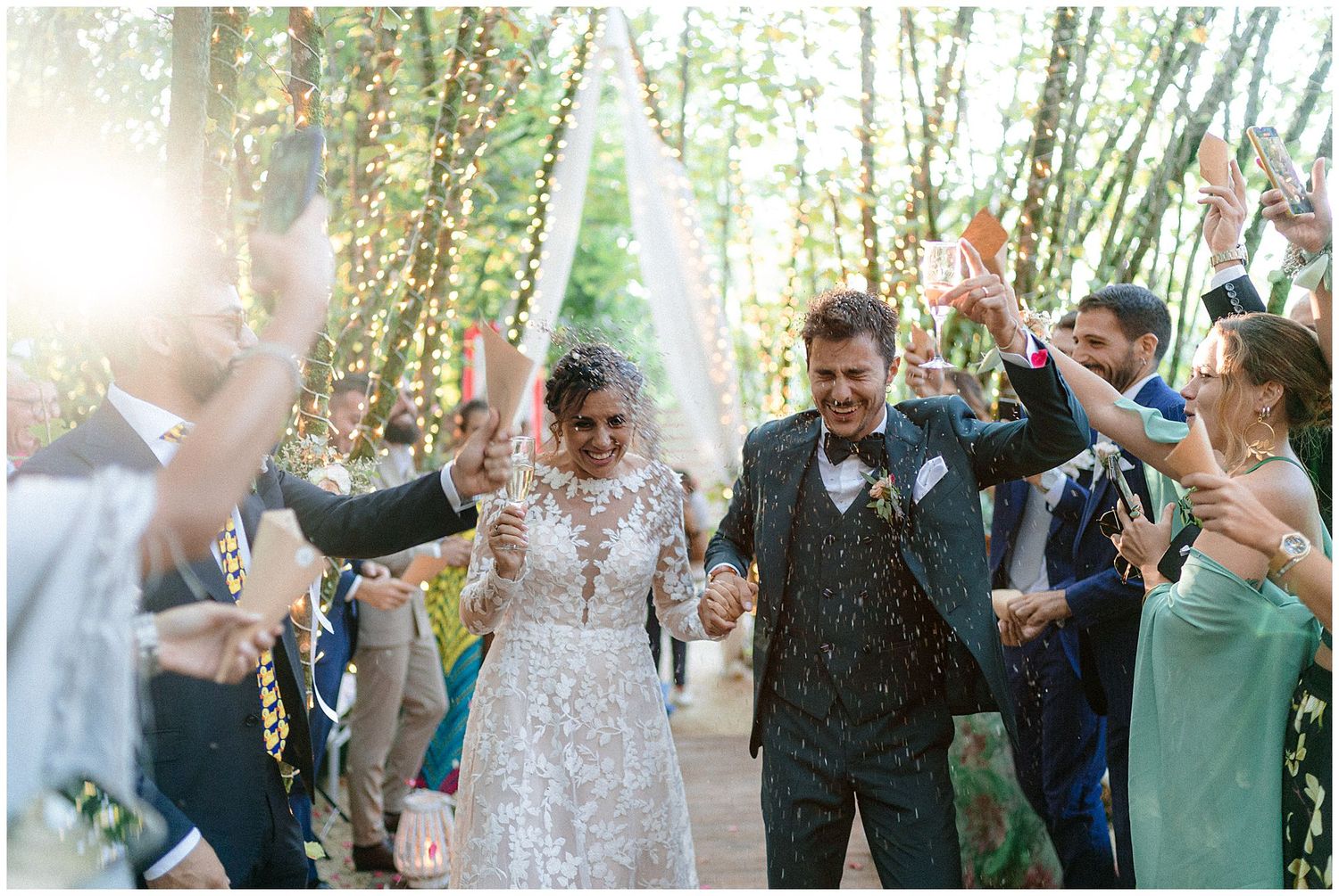 Wedding party celebrating under sunlit trees as festive guests shower newlyweds with confetti.
