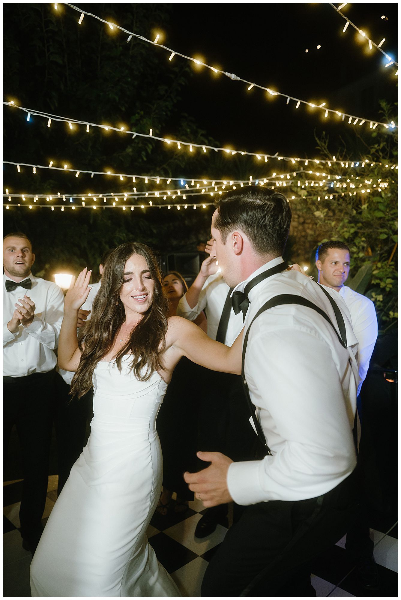 A couple dances together under romantic string lights at an outdoor evening wedding reception celebration.