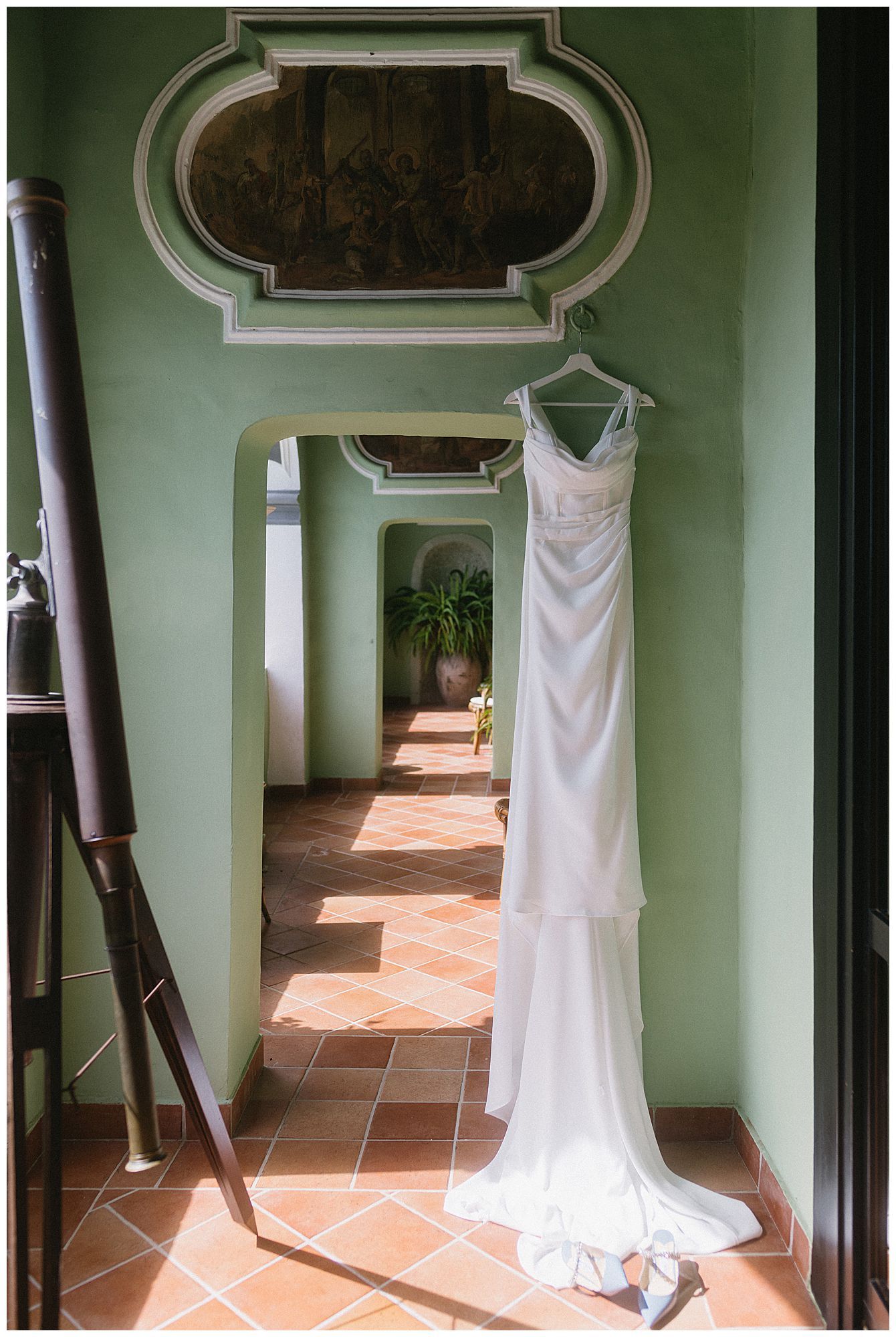 An elegant white wedding dress hangs in a sunlit breezeway with terra cotta floors and mint green walls creating a romantic ambiance.