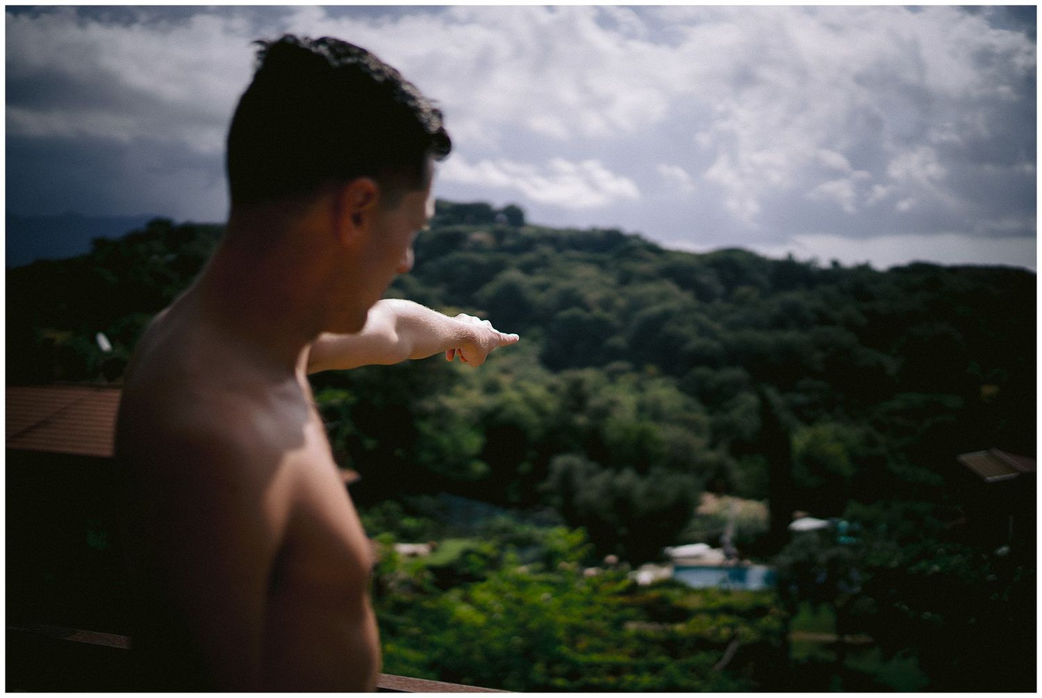 A shirtless person looks out over a lush green tropical hillside on a cloudy day.