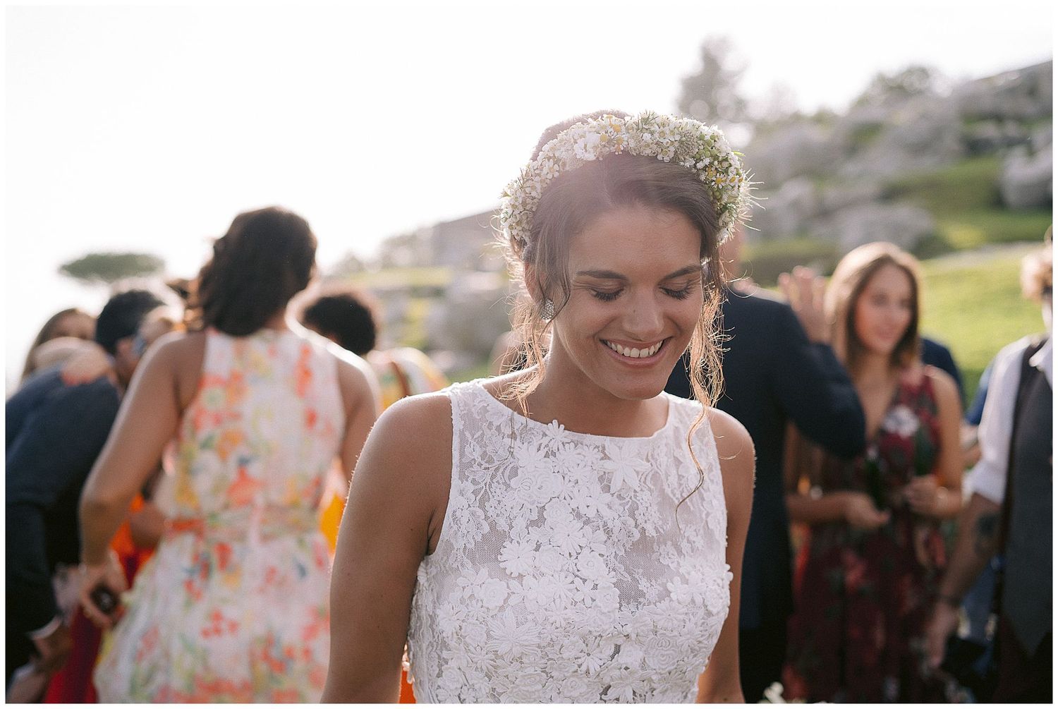 A bride wearing a sleeveless lace wedding dress and tiara smiles outdoors during a wedding celebration surrounded by guests.