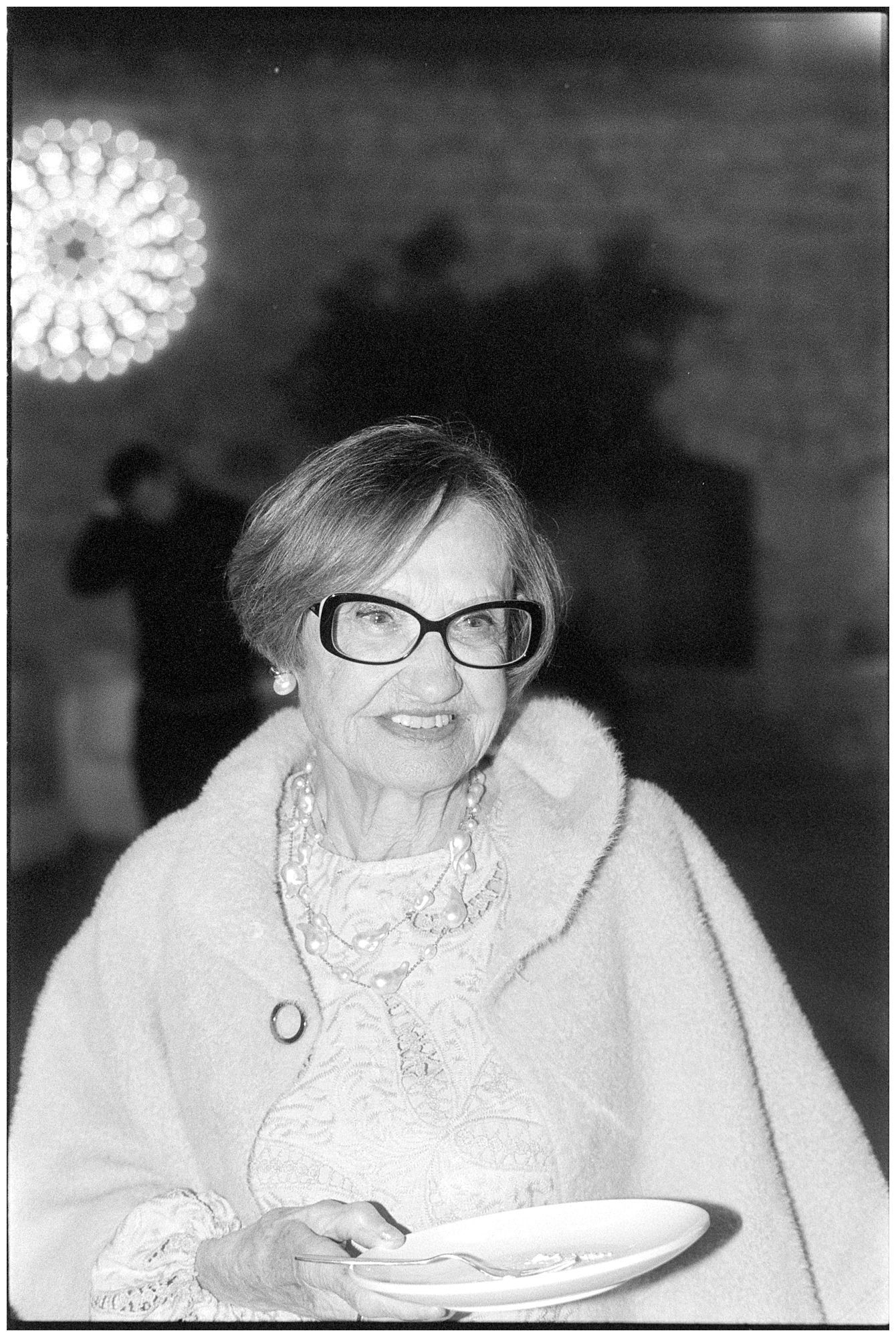 A black and white photograph showing an elderly person in glasses and a white sweater smiling while holding a plate.