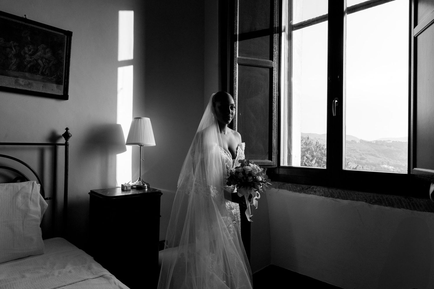 A bride in a white wedding dress and veil stands by a window holding a bouquet in a monochrome room.