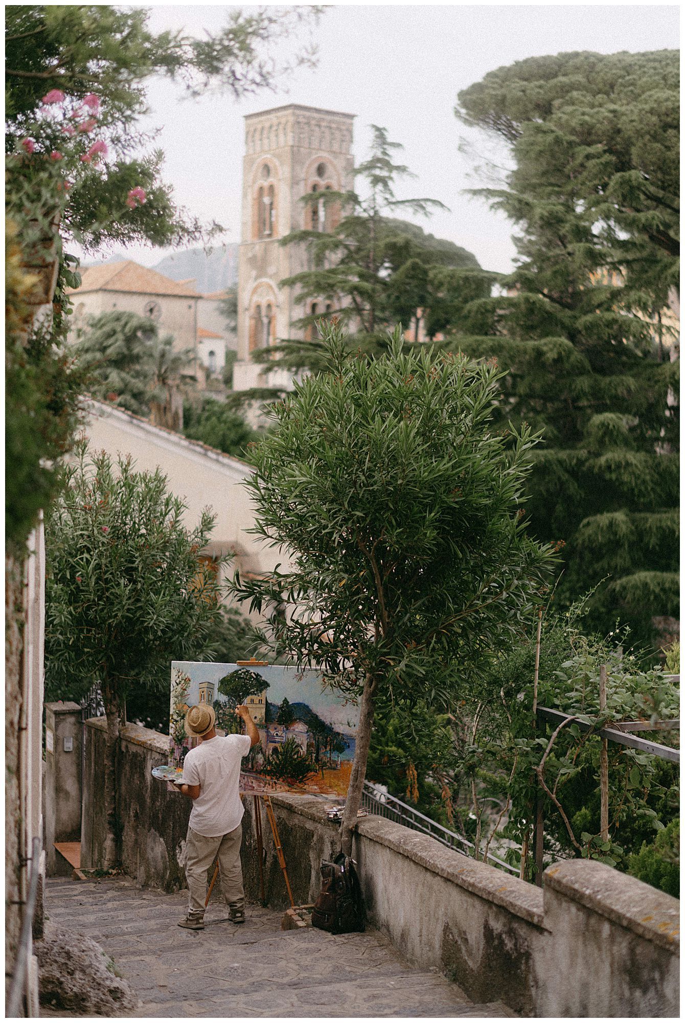 Artist painting on an easel along a picturesque path in Ravello, Italy with a church tower visible in the background.