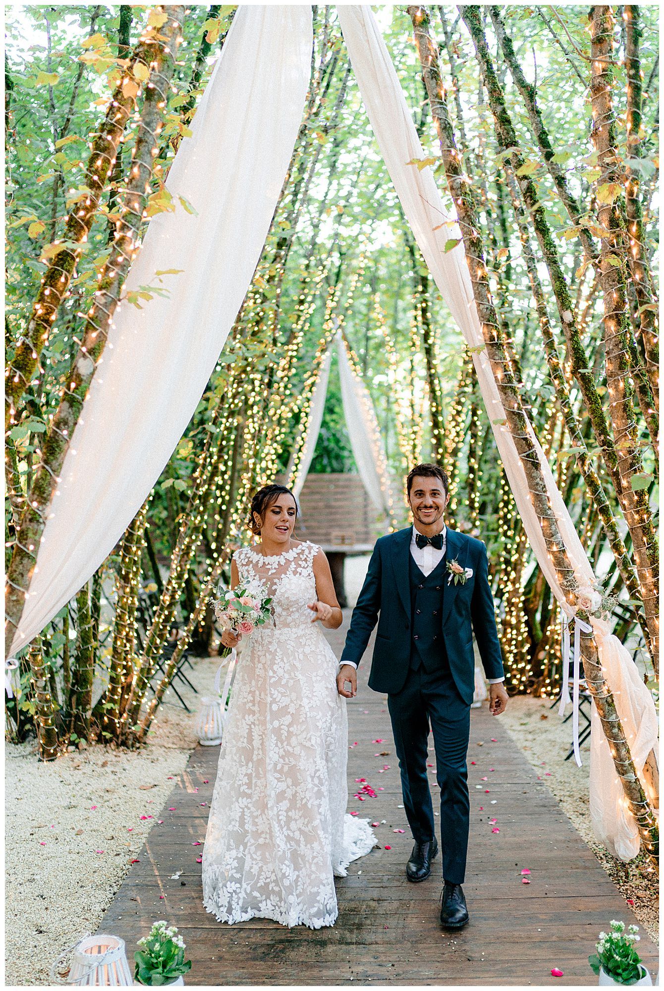 A couple walks down a rustic pathway decorated with white fabric draping and fairy lights among moss-covered trees.