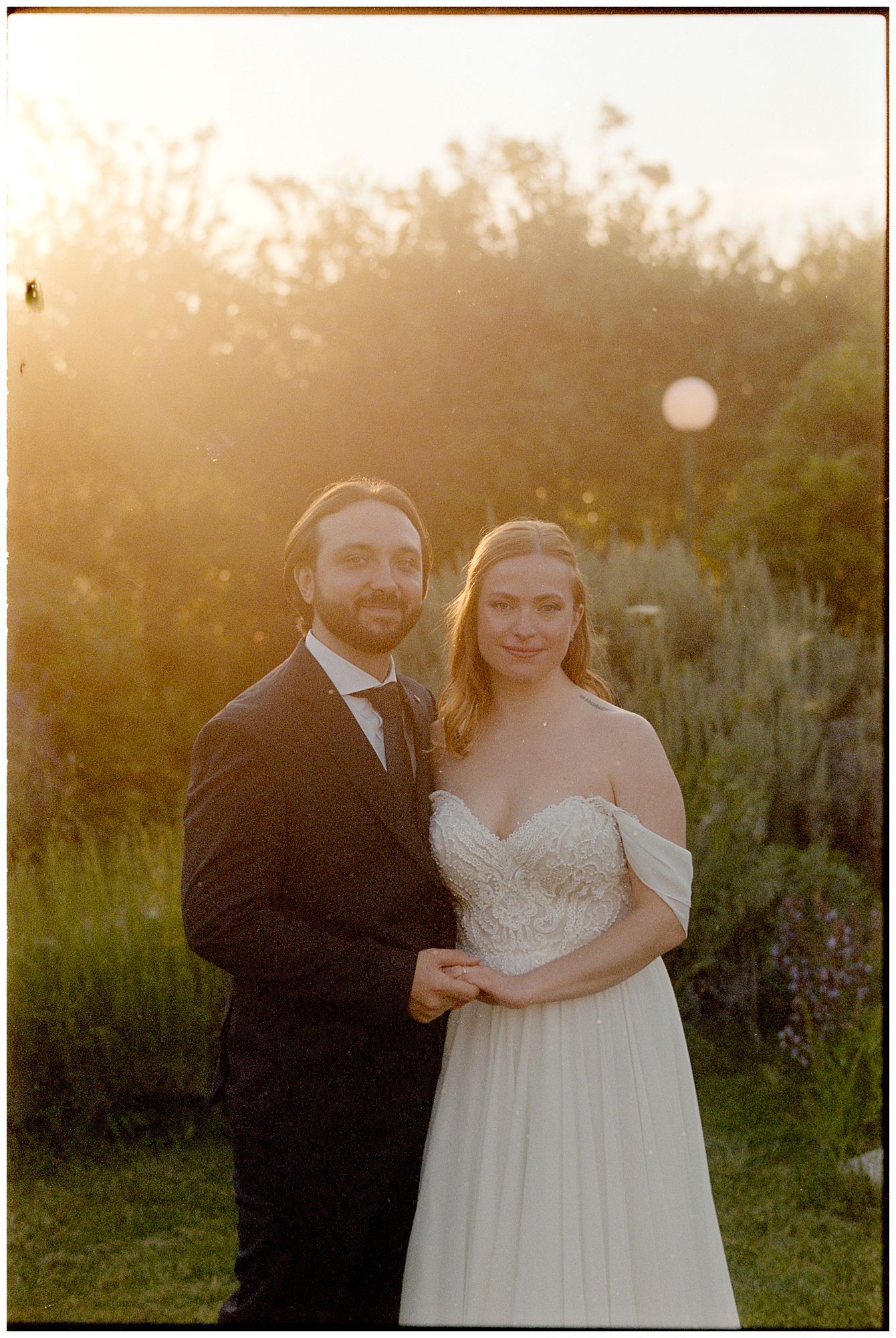 A couple poses in formal wedding attire during golden hour with sunlight and greenery in the background.
