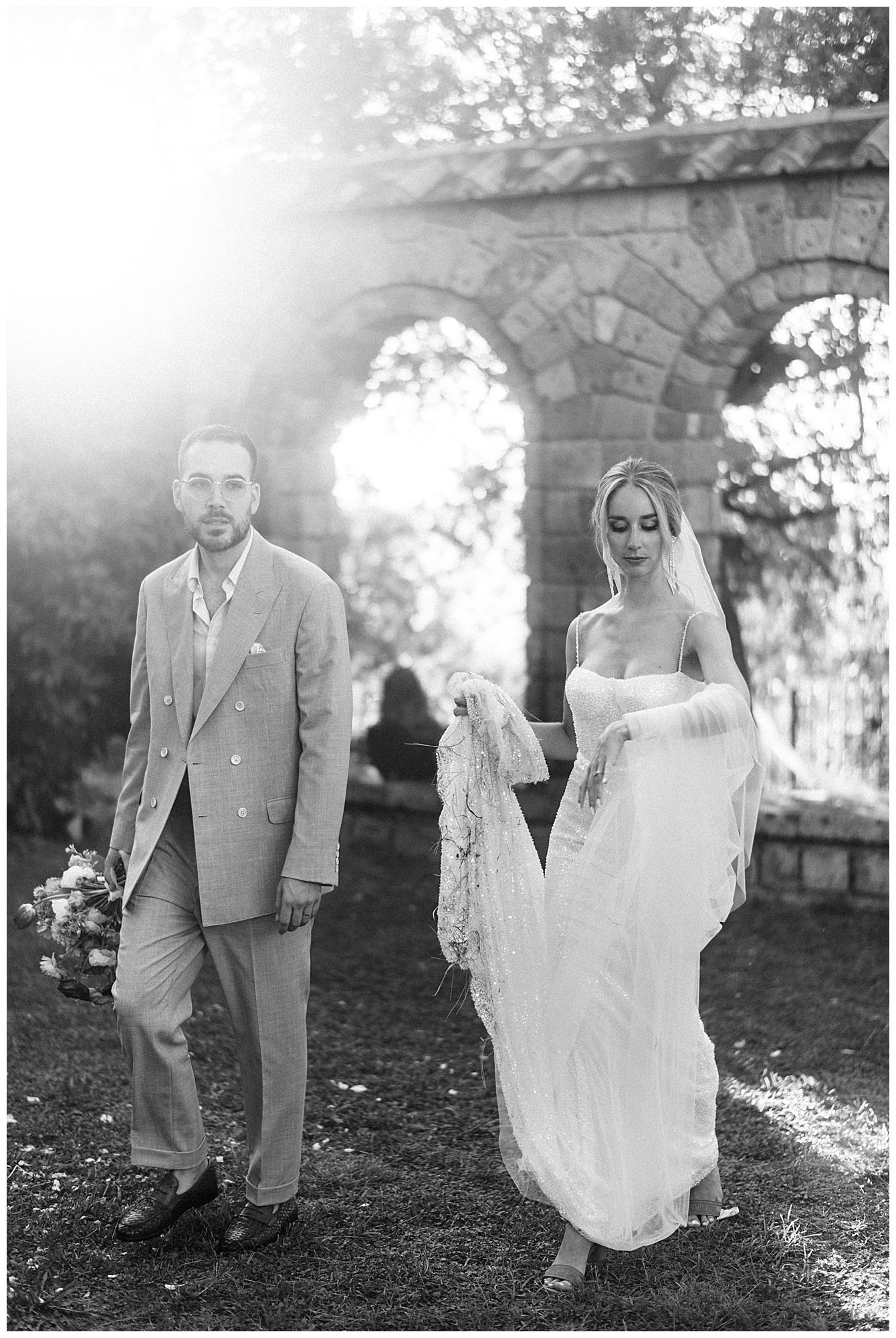 A couple walks together outdoors on their wedding day with stone archways in the background.