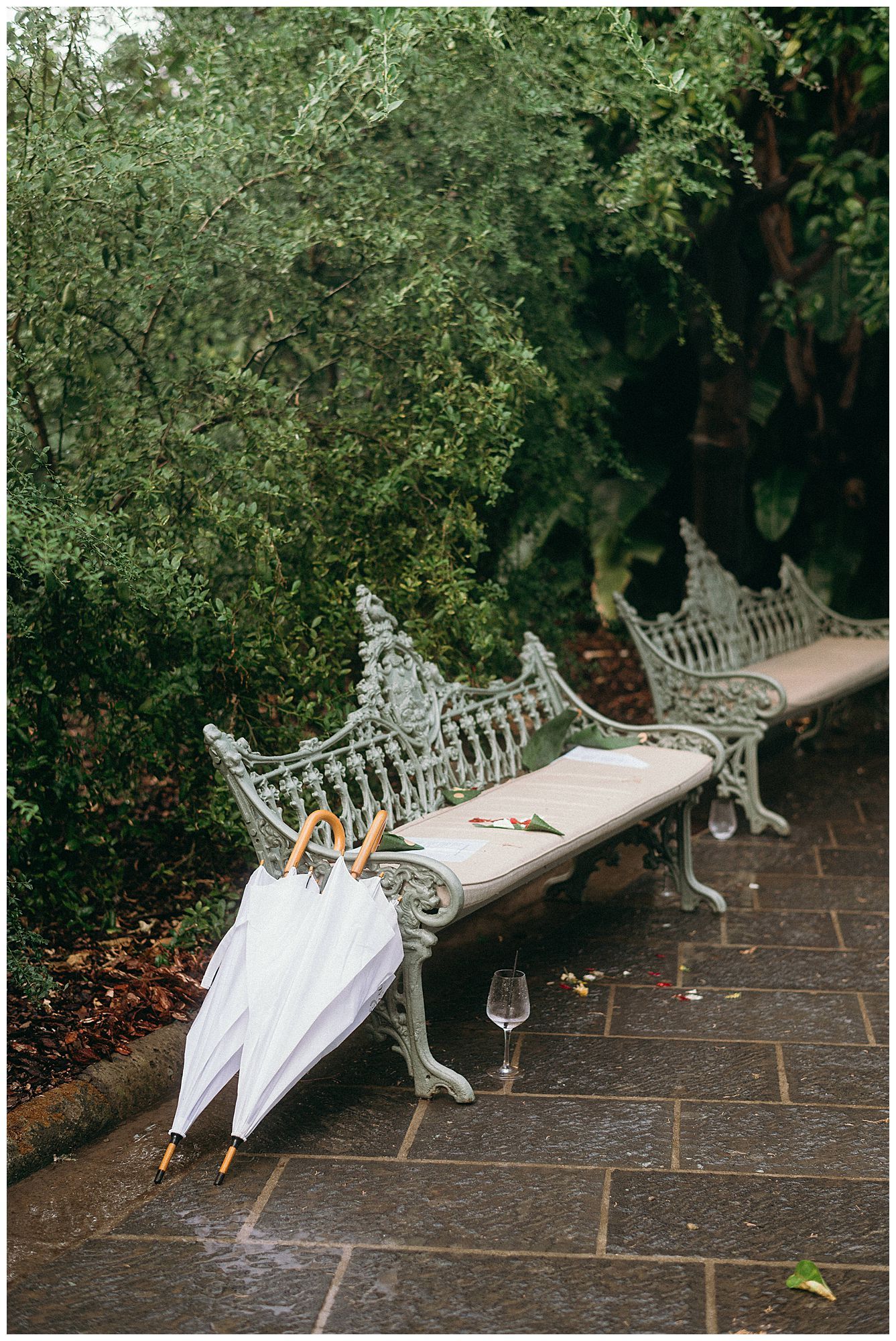 White wedding umbrellas resting on an ornate vintage cast iron garden bench along a stone patio surrounded by greenery.