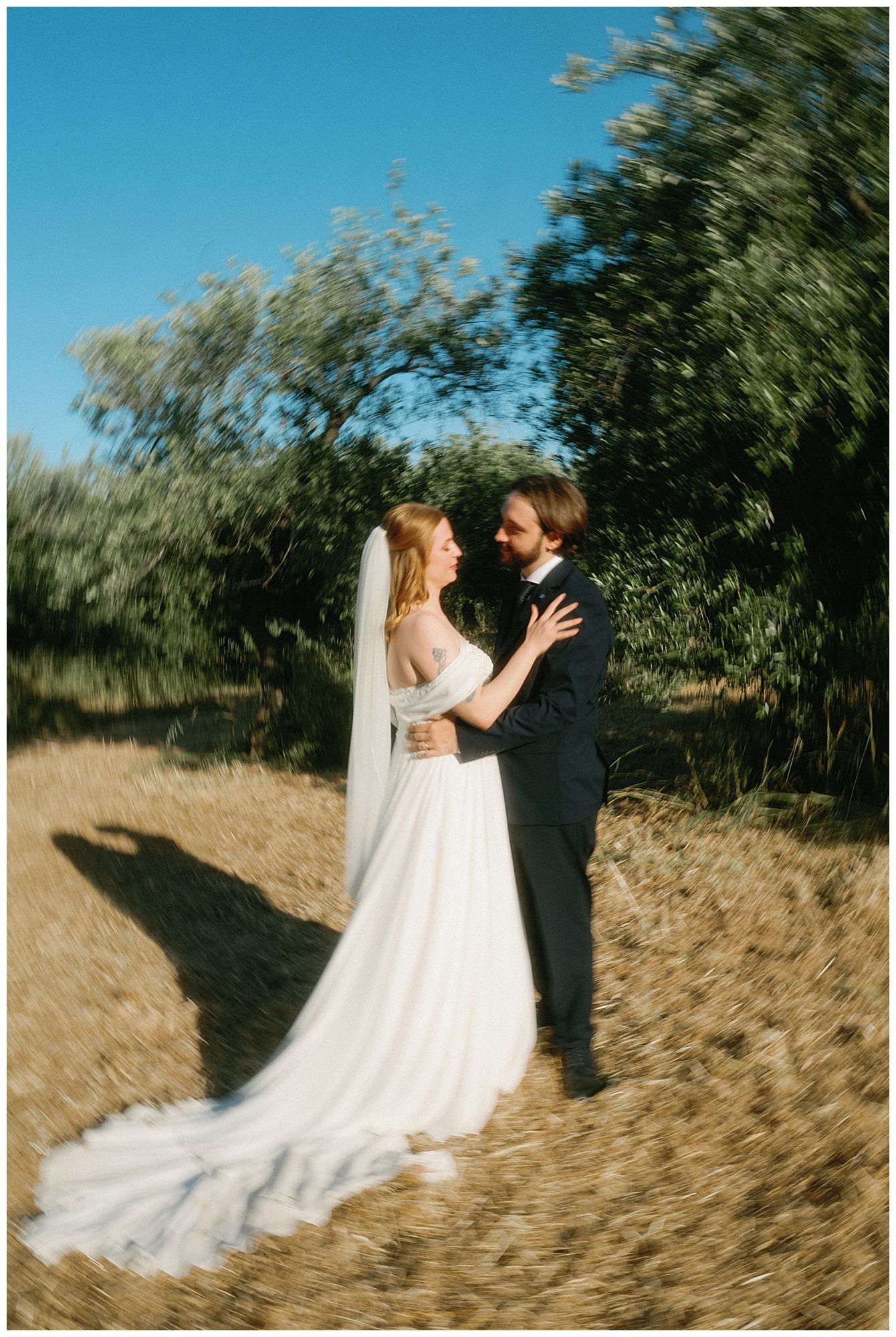 A couple embraces outdoors in wedding attire with a flowing white dress and veil against green foliage and blue sky.