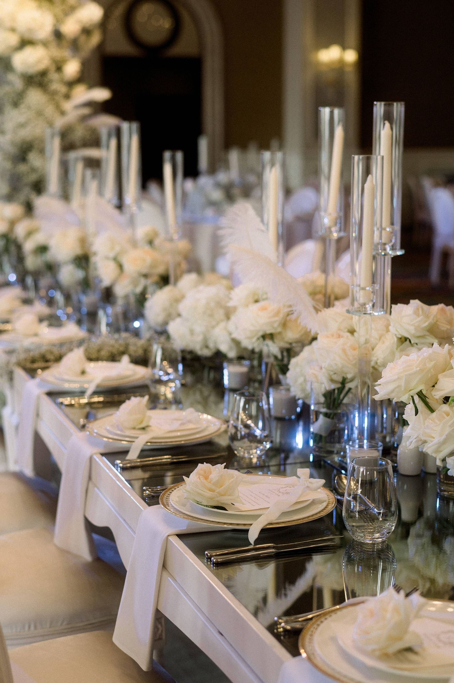 Elegant wedding table setting with white floral centerpieces, tall glass candle holders, and formal place settings at a reception.