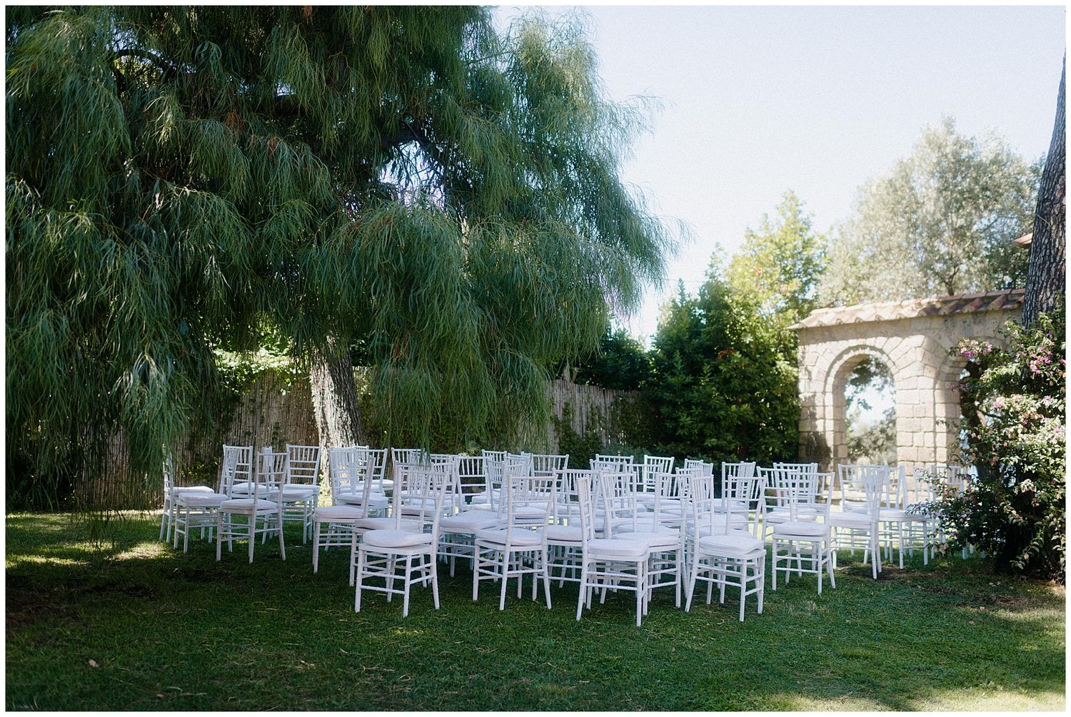 White wedding chairs arranged on a lawn under weeping willow trees with a stone arch in the background for an outdoor ceremony.