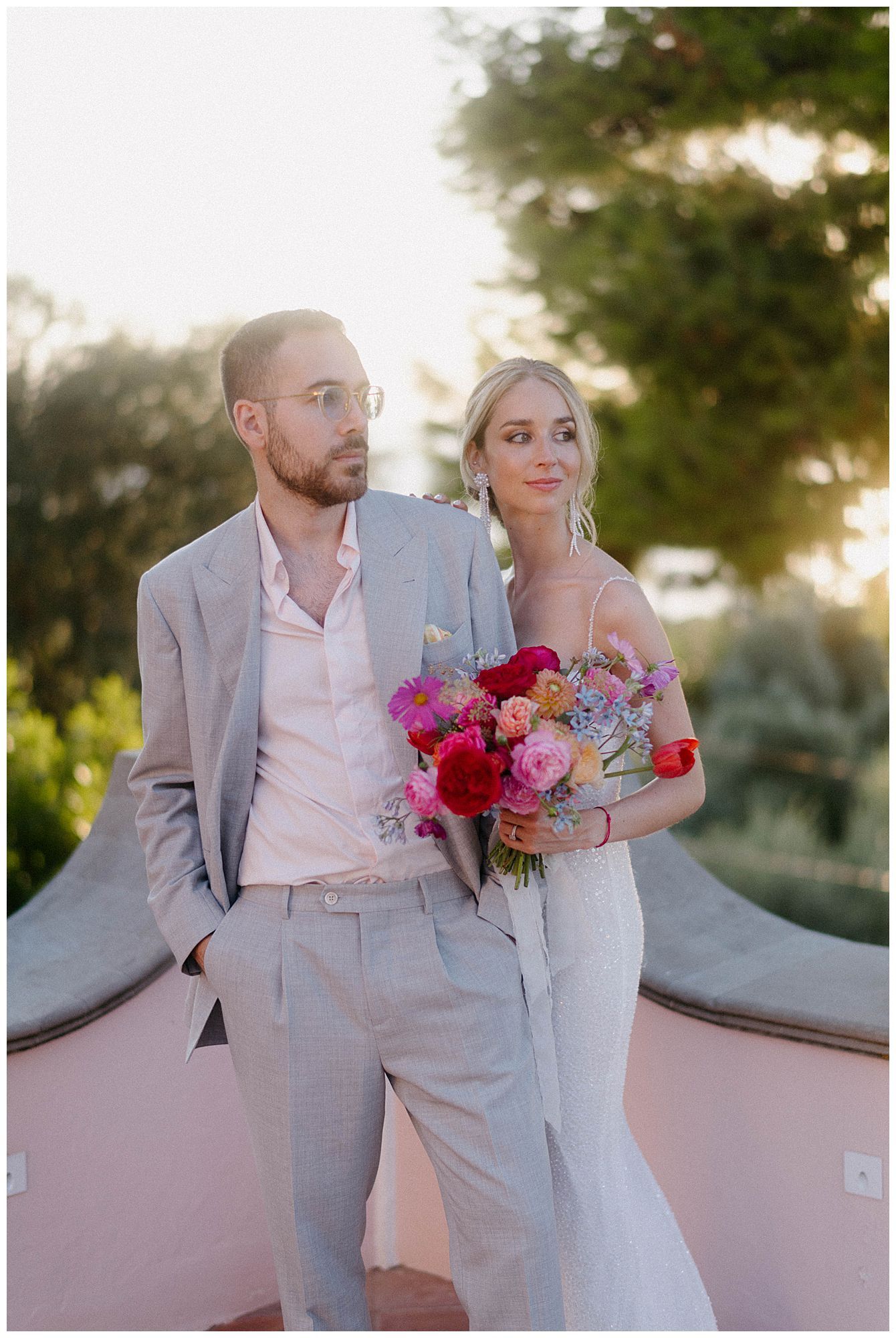 A couple poses together at sunset, the bride holding a vibrant bouquet of pink and red flowers against a garden backdrop.