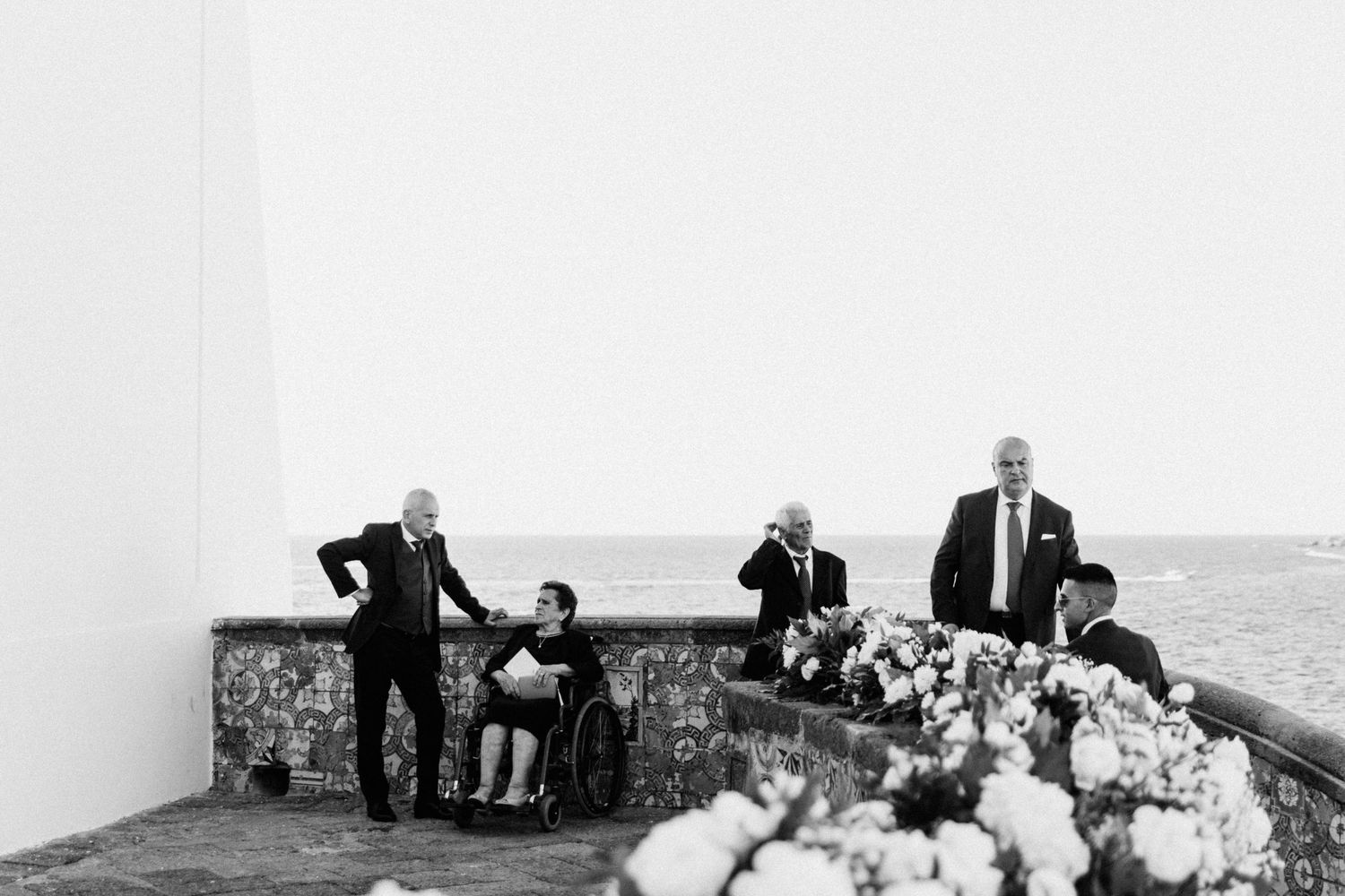 A black and white photo captures wedding guests standing on a stone terrace overlooking a scenic vista.