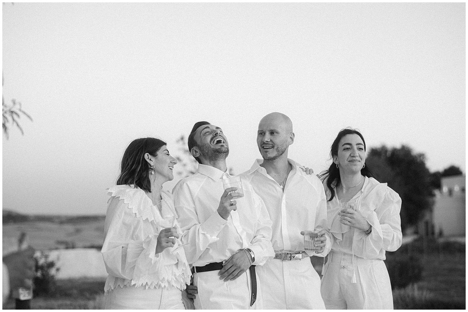 Group of wedding guests in white attire laughing together during outdoor celebration.