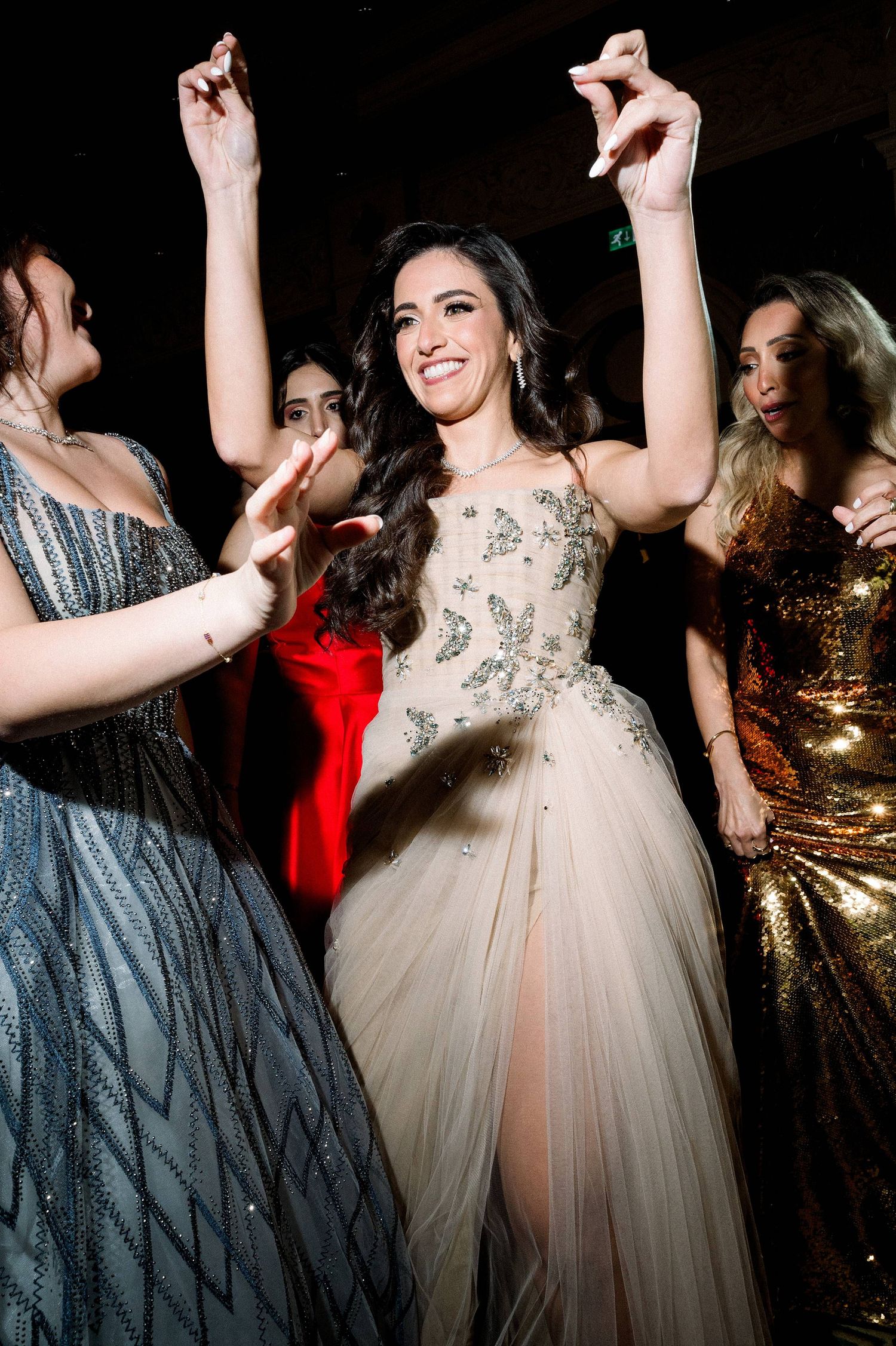 Elegant woman in beaded tulle gown dances with raised arms at formal party, surrounded by guests in metallic dresses.