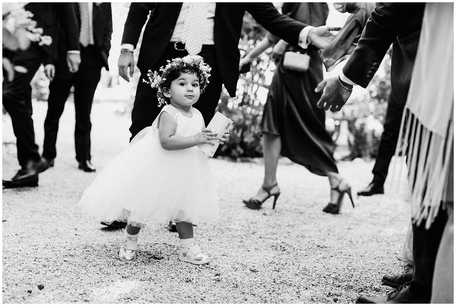 A black and white film photograph of wedding guests' legs and shoes gathered on a gravel path.