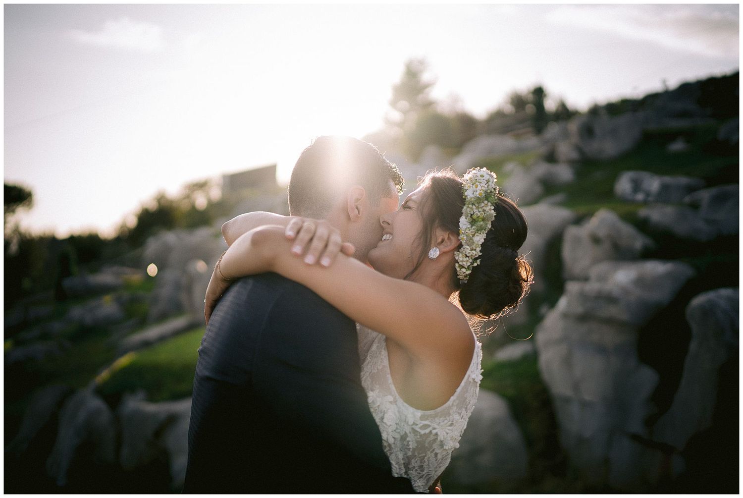 Two people embrace in a romantic moment at sunset among rocky terrain with lens flare highlighting the scene.