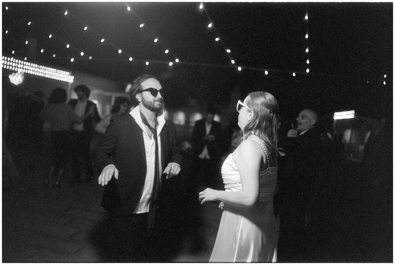 Two people dancing and laughing together at a nighttime celebration with string lights overhead.