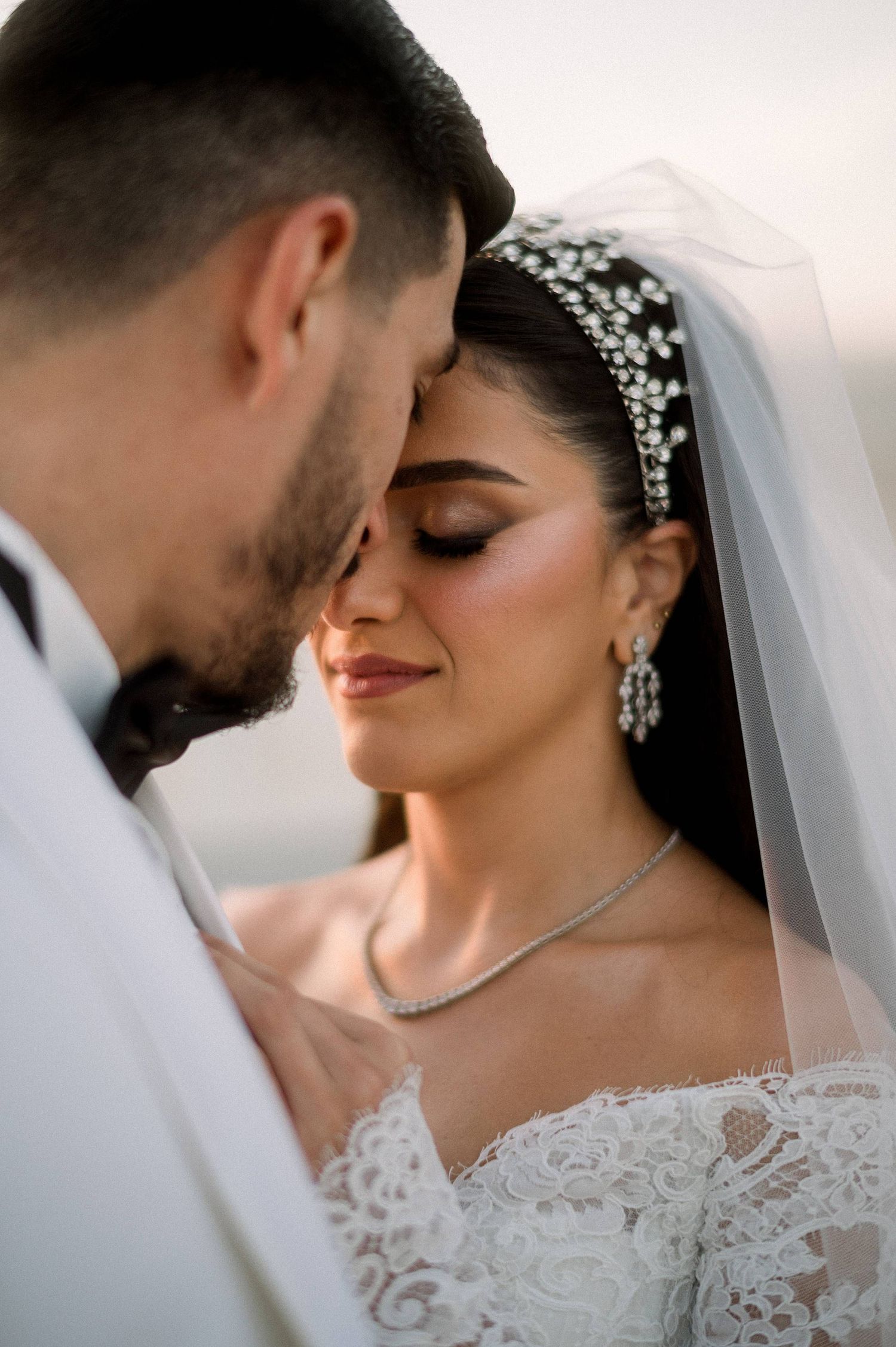 A tender wedding moment with a bride wearing lace, crystal headpiece and veil alongside groom in a white suit.