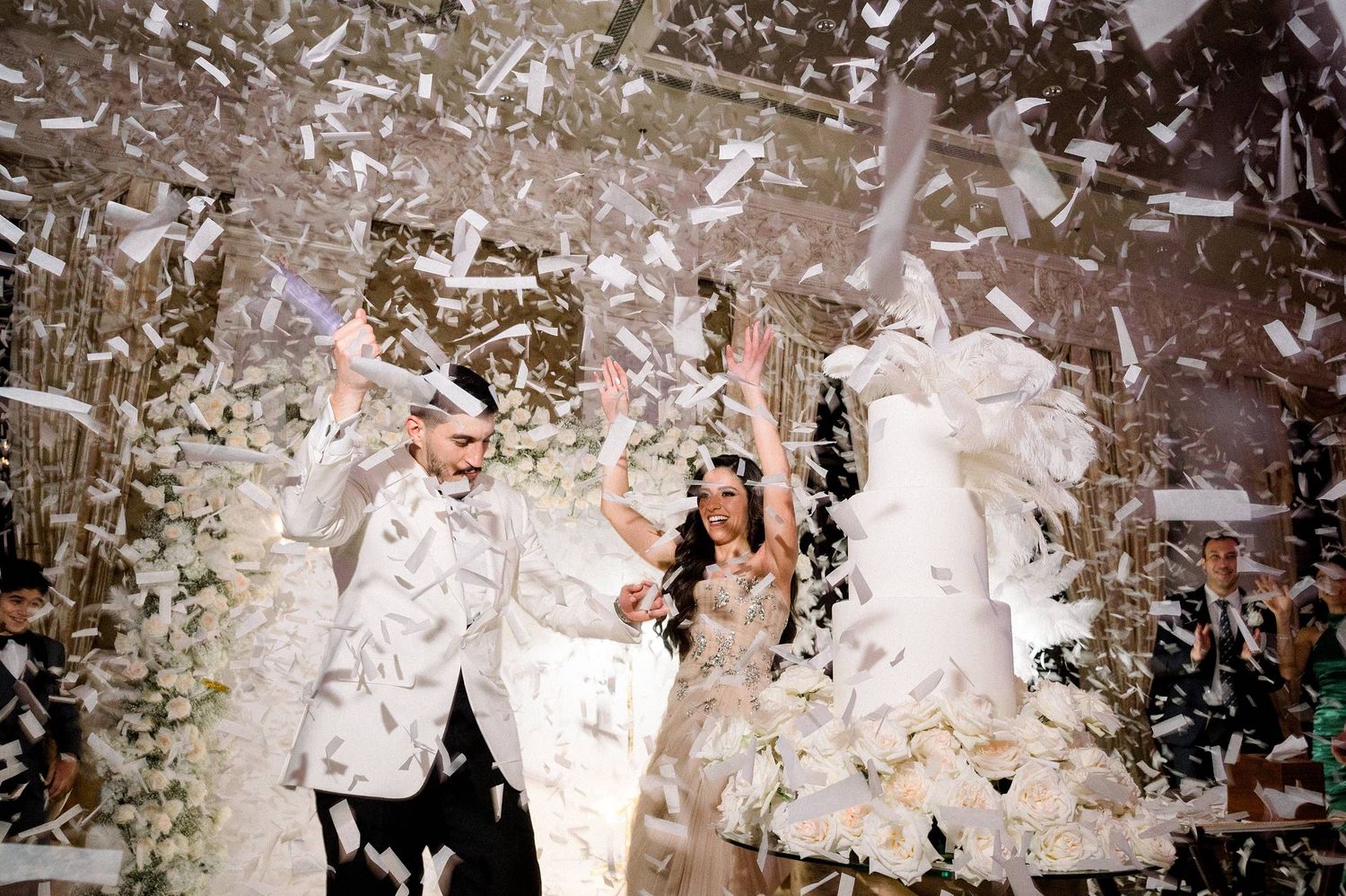 Happy couple celebrates their wedding reception under a cascade of white confetti in a joyous party atmosphere.