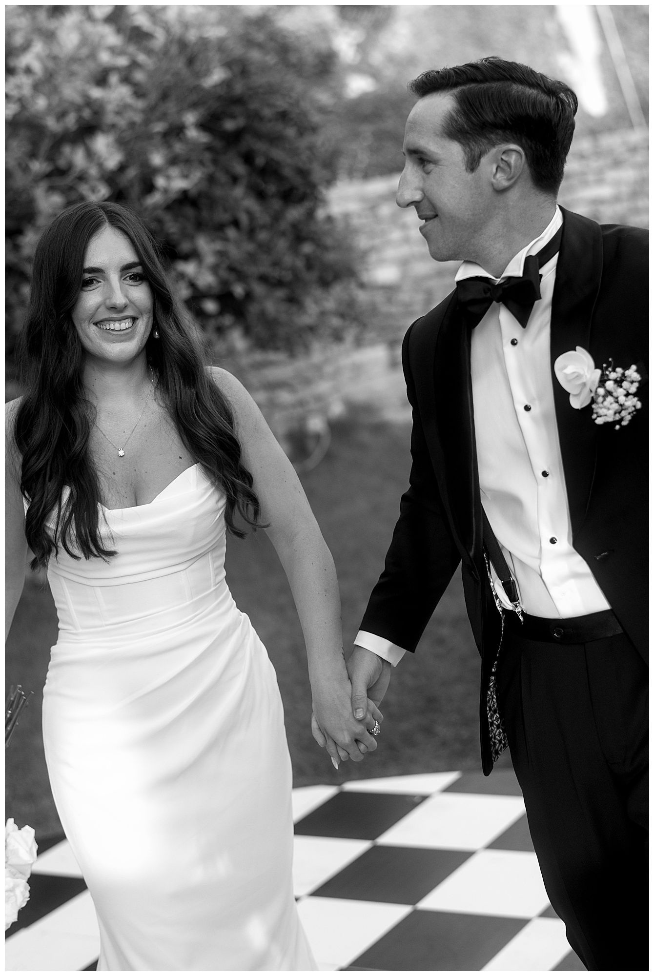 Black and white wedding photo of newlyweds joyfully walking together on a checkered floor outdoors.