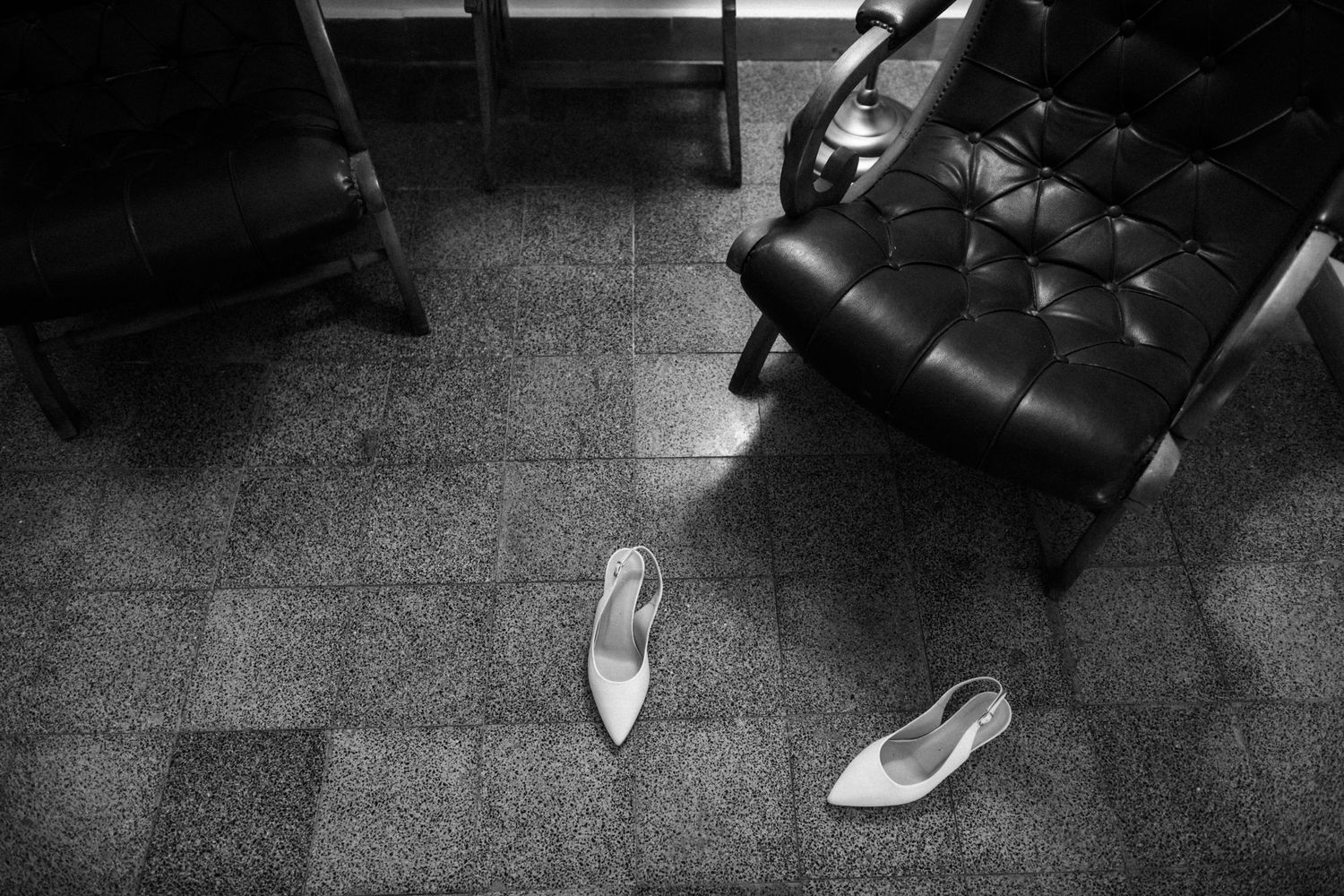A black and white photograph shows a pair of white ballet flats on a dark tiled floor next to a leather chair.