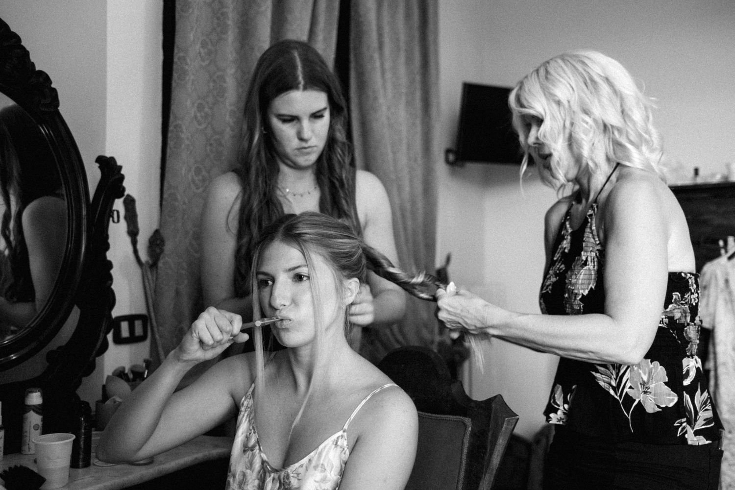 Black and white photo of a wedding party getting ready, with stylists working on hair and makeup preparations.
