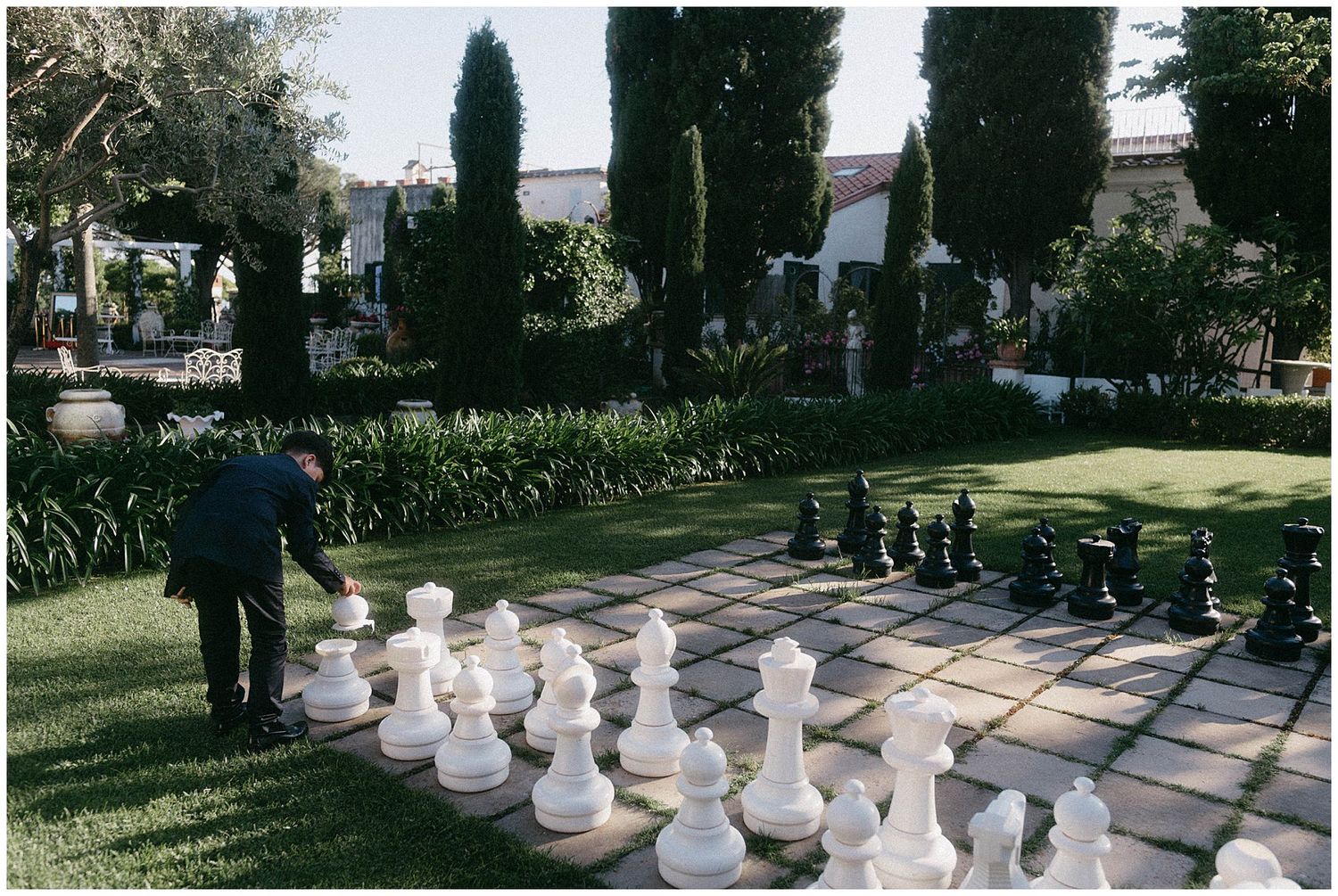 Giant chess pieces on a paved garden path with tall cypress trees and landscaping in the background.