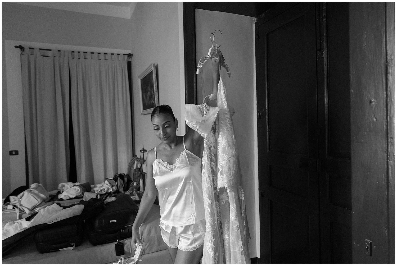 Black and white photo showing someone in a white top next to a hanging wedding dress in a bedroom setting.