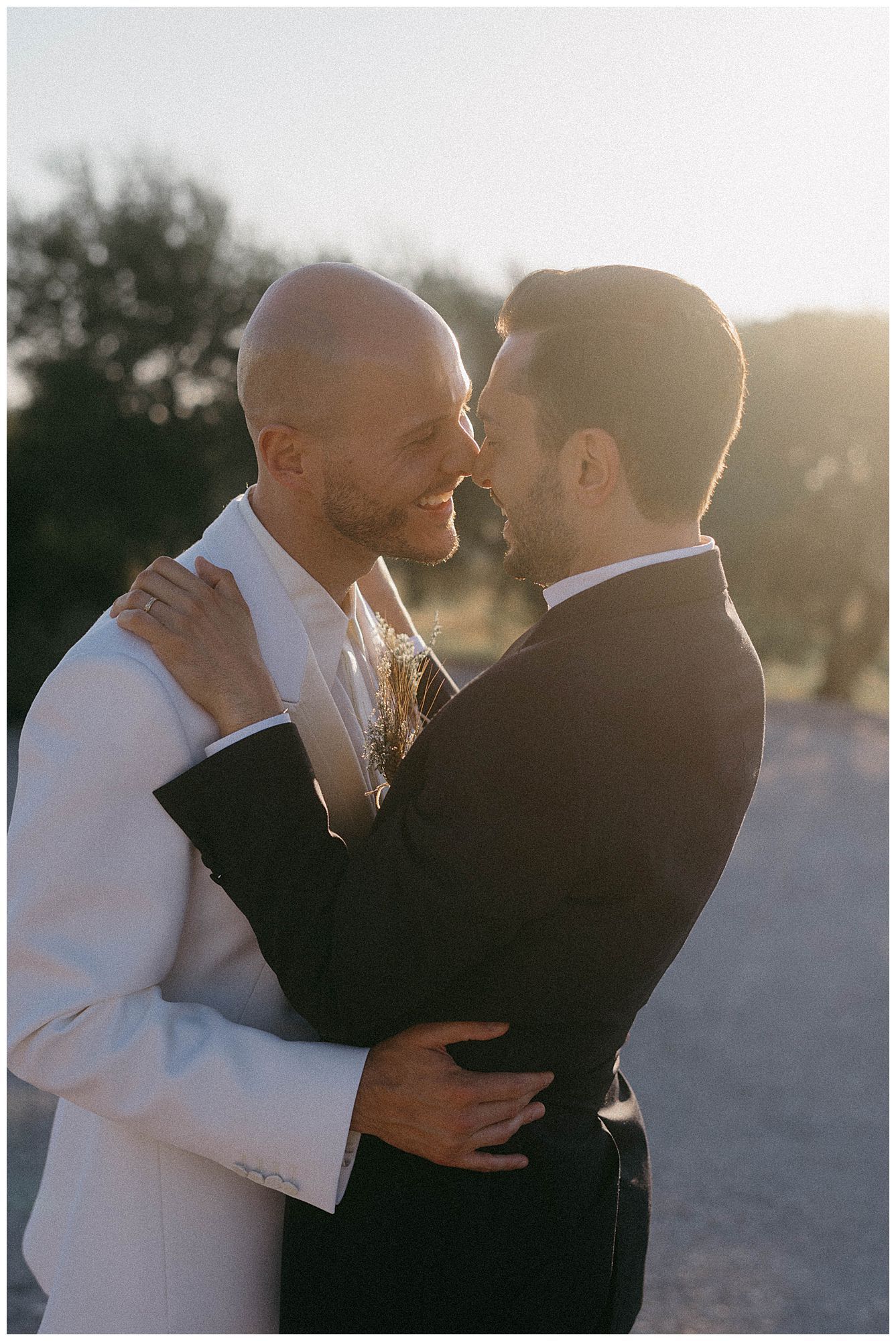 A couple in wedding attire share an intimate moment at sunset, with one wearing white and one in black.