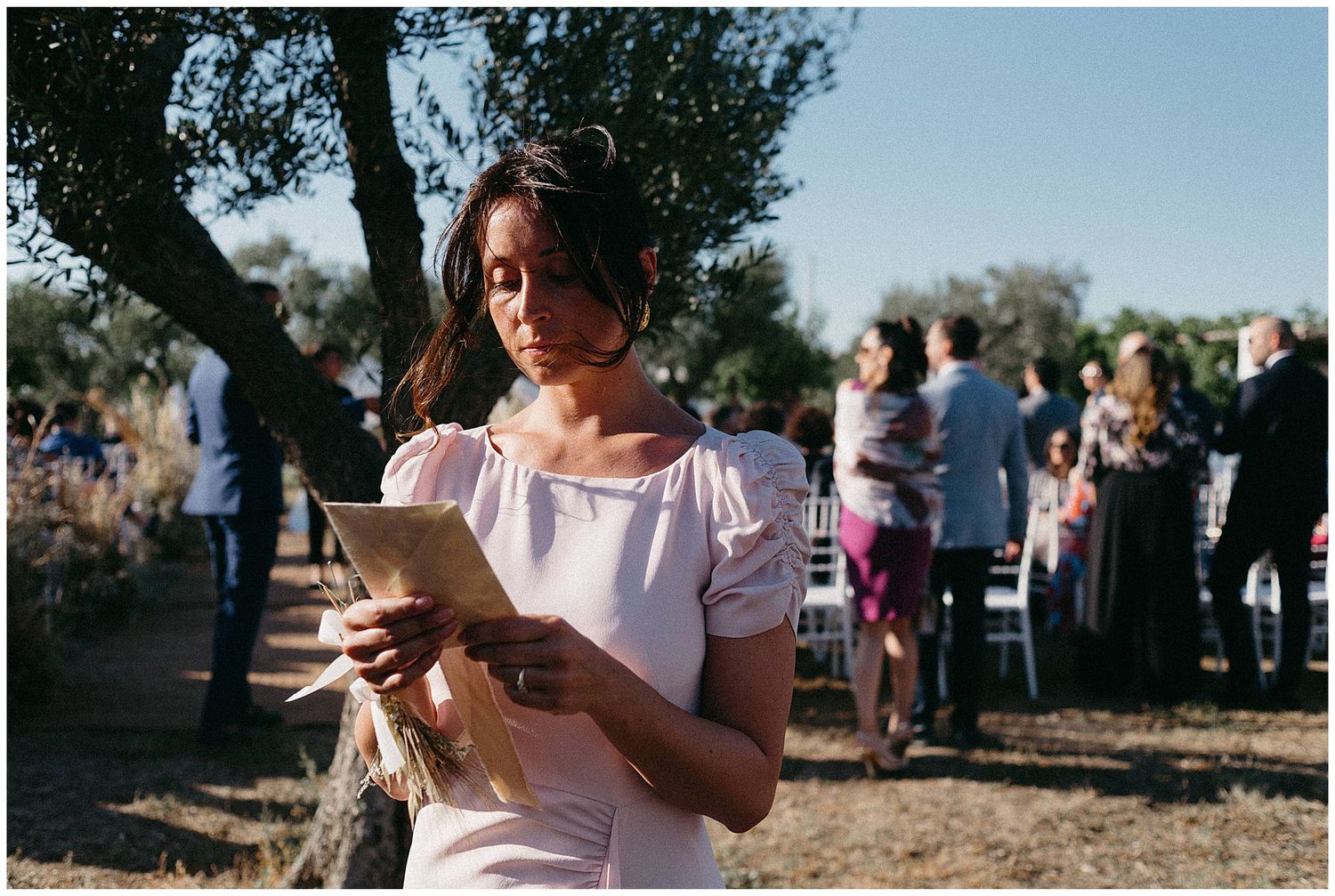 Person in white dress checks phone at outdoor gathering with crowd and trees in background during daytime event.