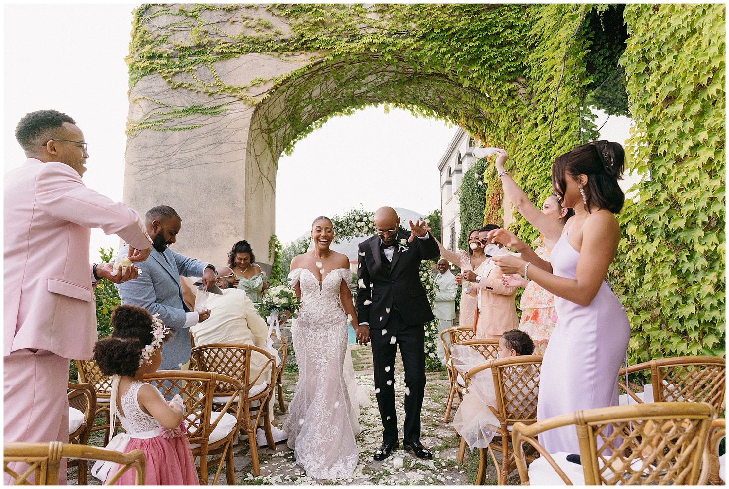 Wedding party celebrating with confetti in an ivy-covered archway during outdoor garden ceremony.