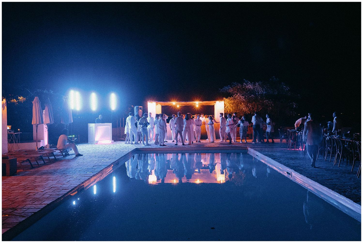 People gathered around an illuminated swimming pool at night with bright lights and lounge chairs by the poolside.