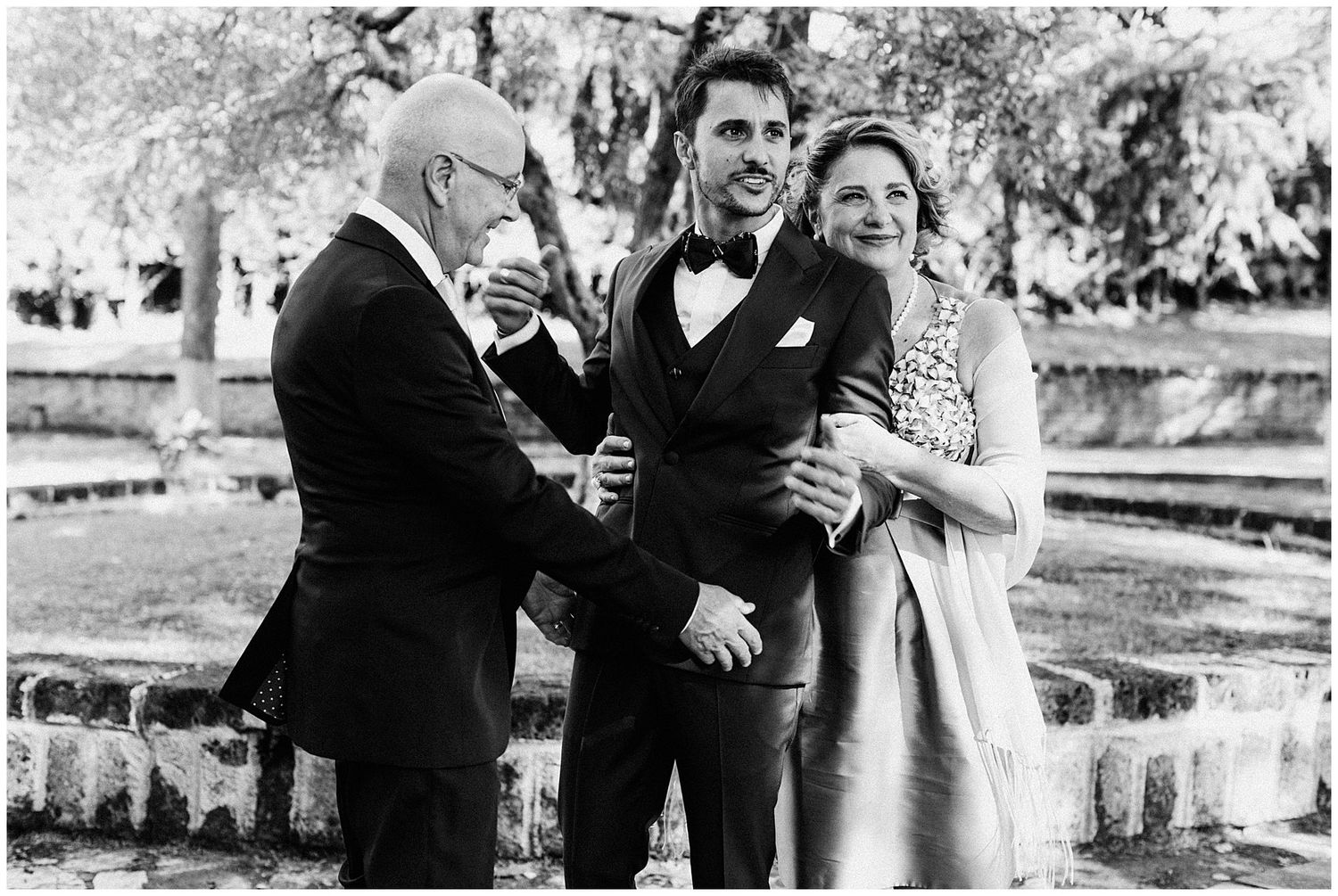A black and white panoramic wedding photo showing couples enjoying candid moments by a scenic lakeside garden.