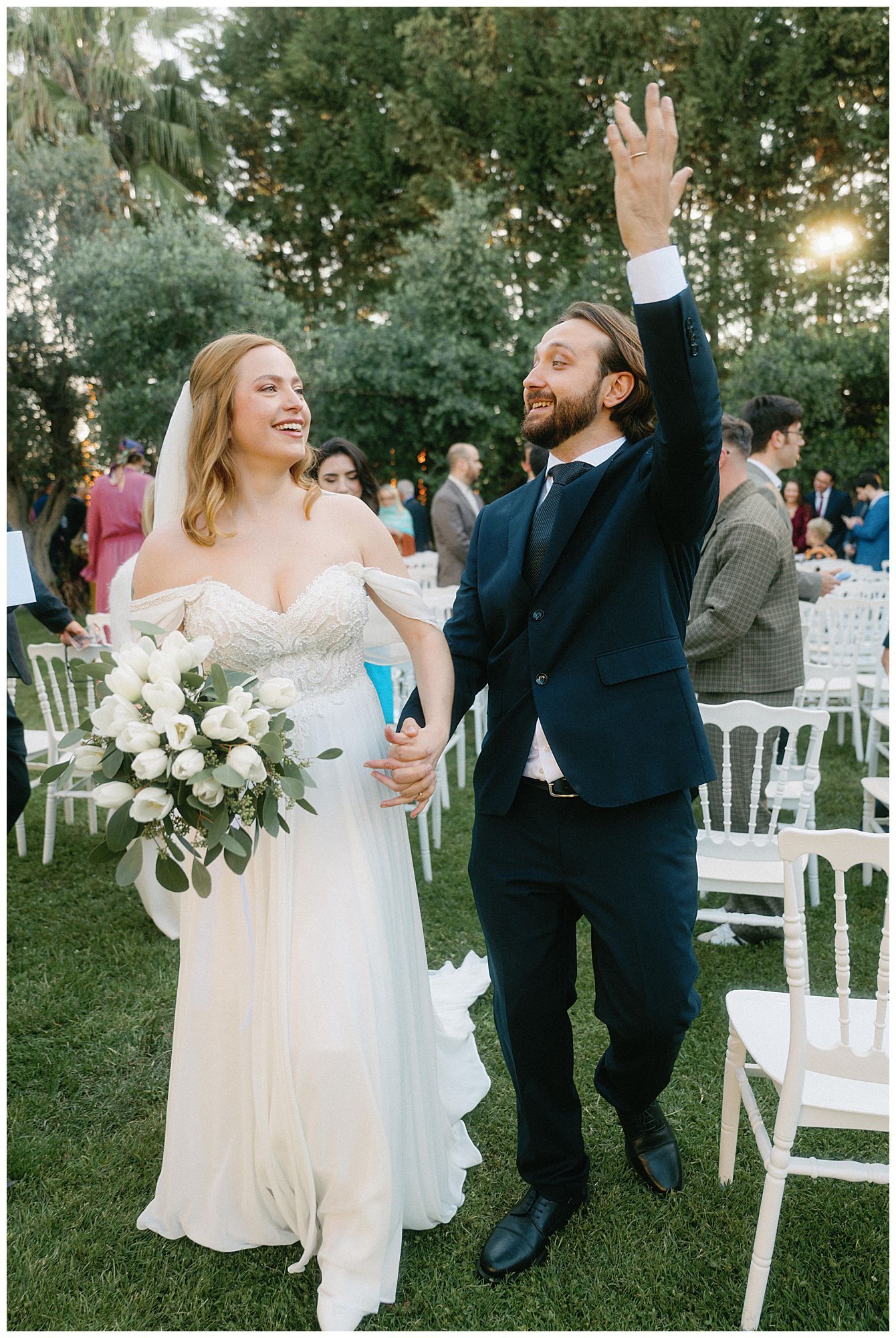 A joyful wedding celebration outdoors with a couple surrounded by white chairs and garden landscaping at sunset.