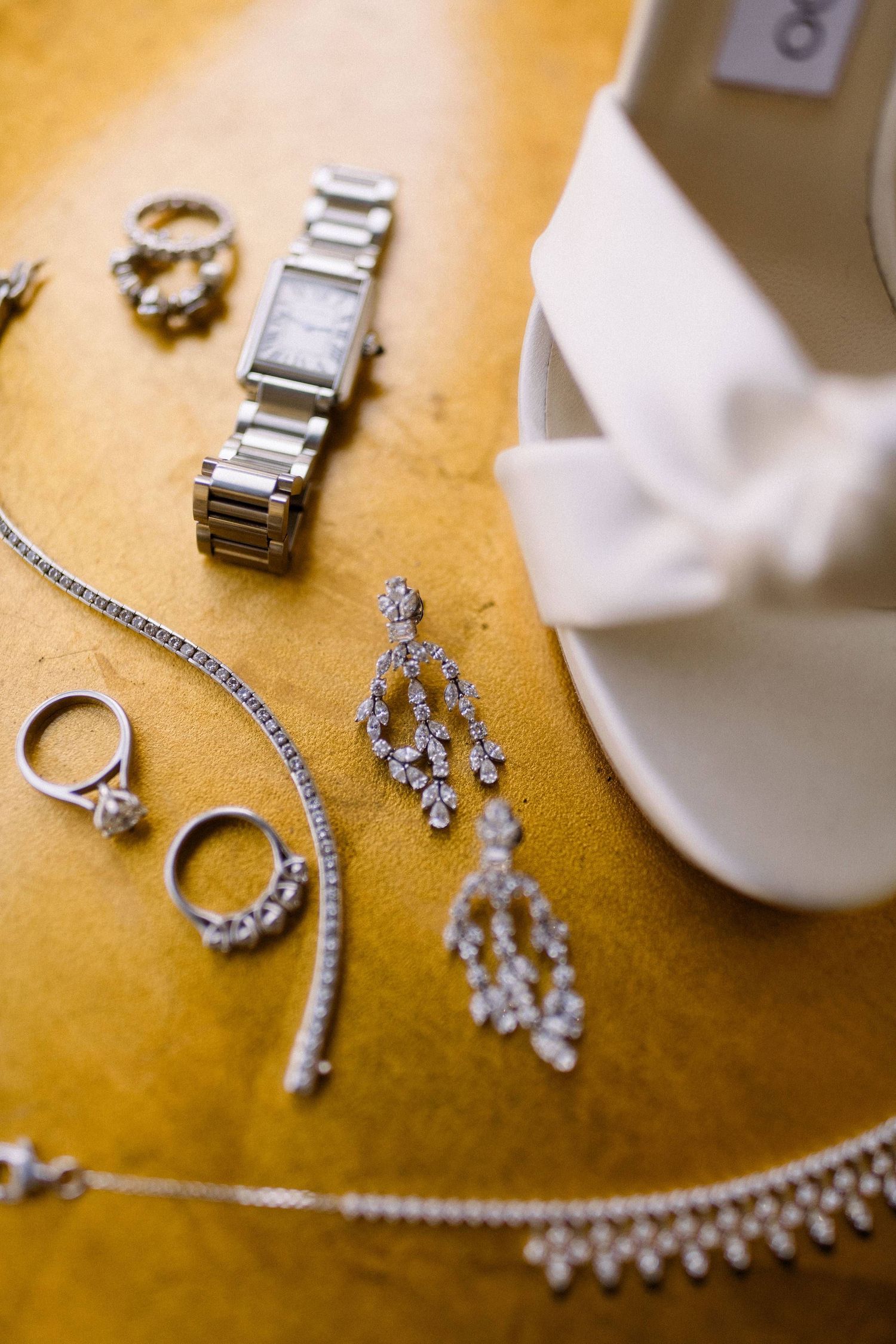 Wedding accessories including sparkly diamond jewelry, silver rings, chandelier earrings and a watch laid out on a gold surface.
