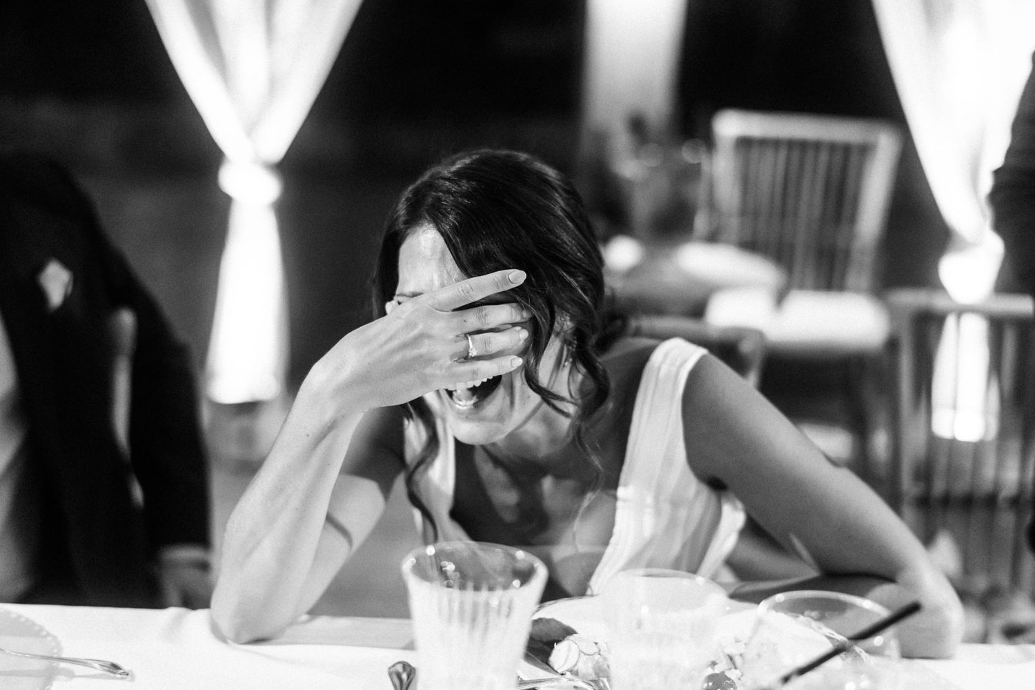 Black and white photo of someone covering their face while seated at a table with white curtains in the background.