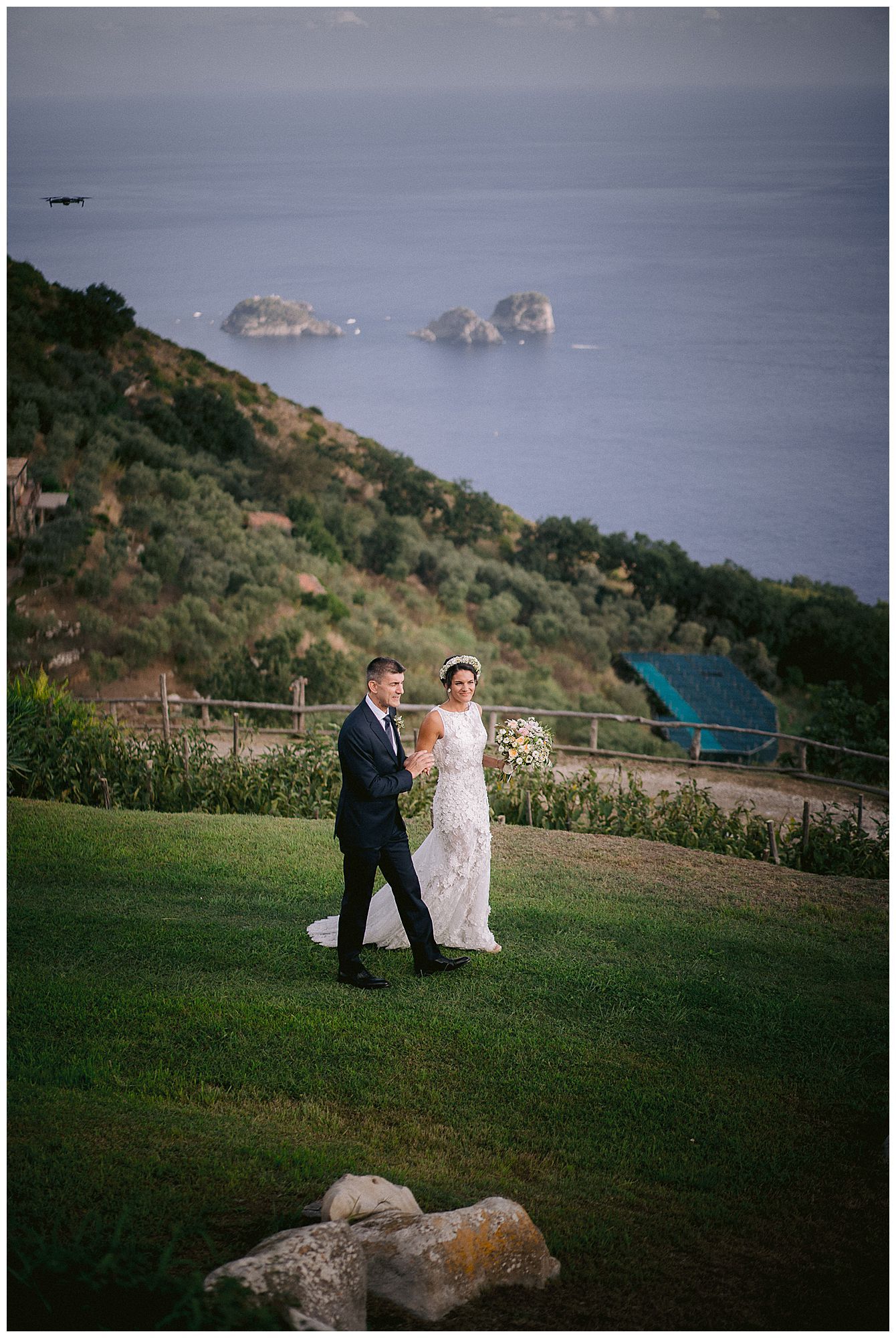 A couple in formal attire walk along a grassy hillside overlooking rocky ocean cliffs on a misty coastal afternoon.