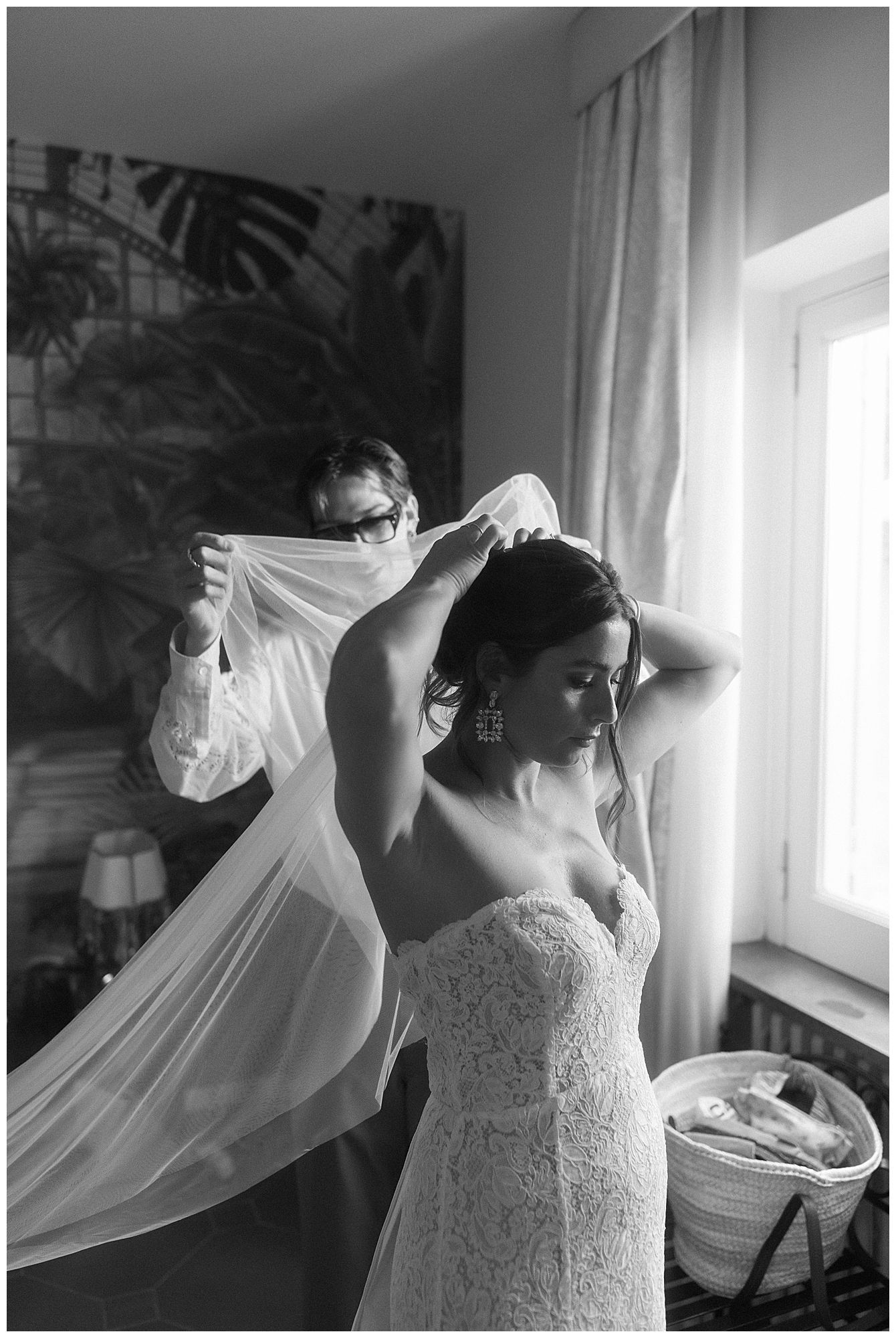 Black and white photo capturing a tender moment during wedding preparations as a veil is delicately placed near a window.