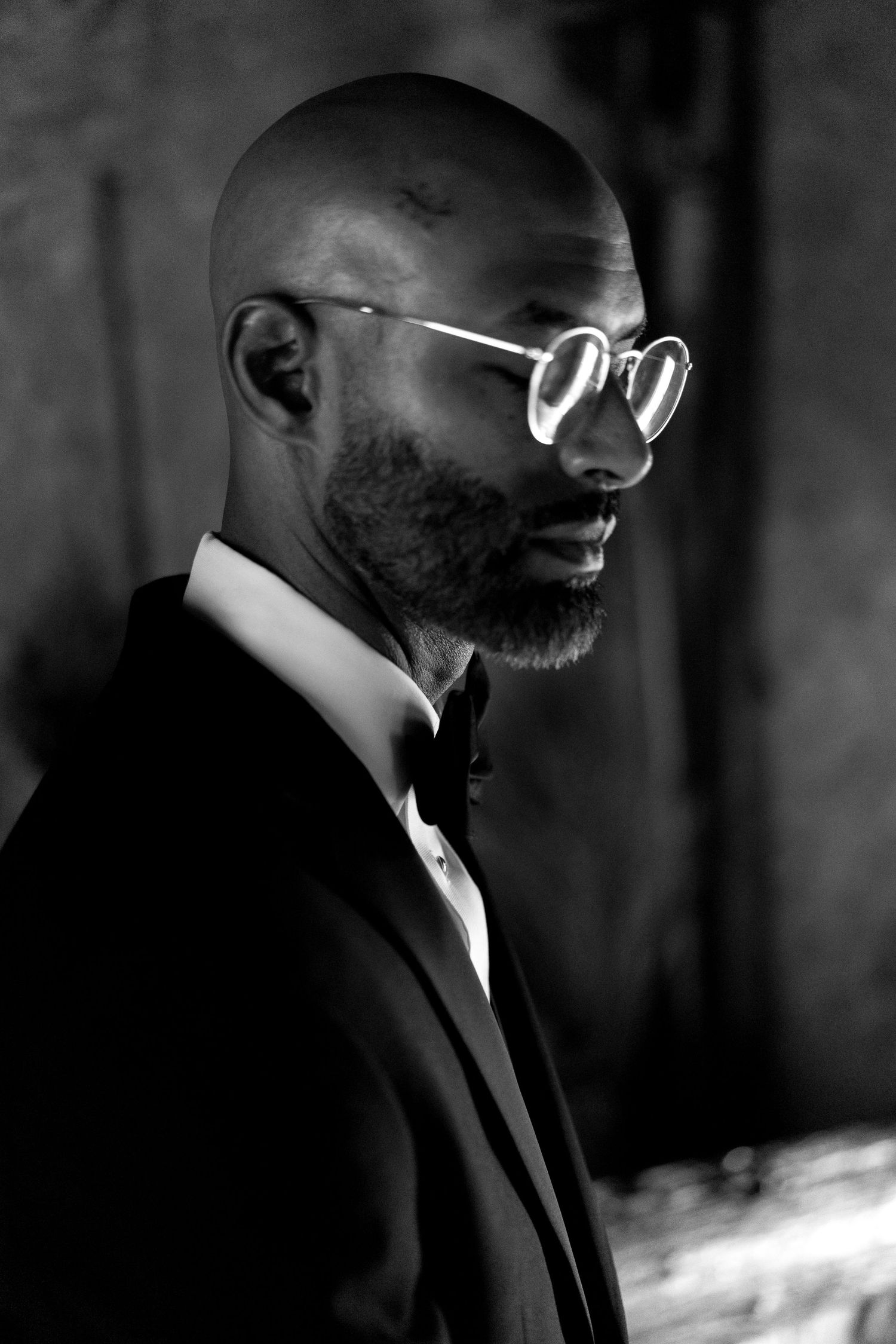 Artistic black and white portrait of person in tuxedo and round glasses against dark background.
