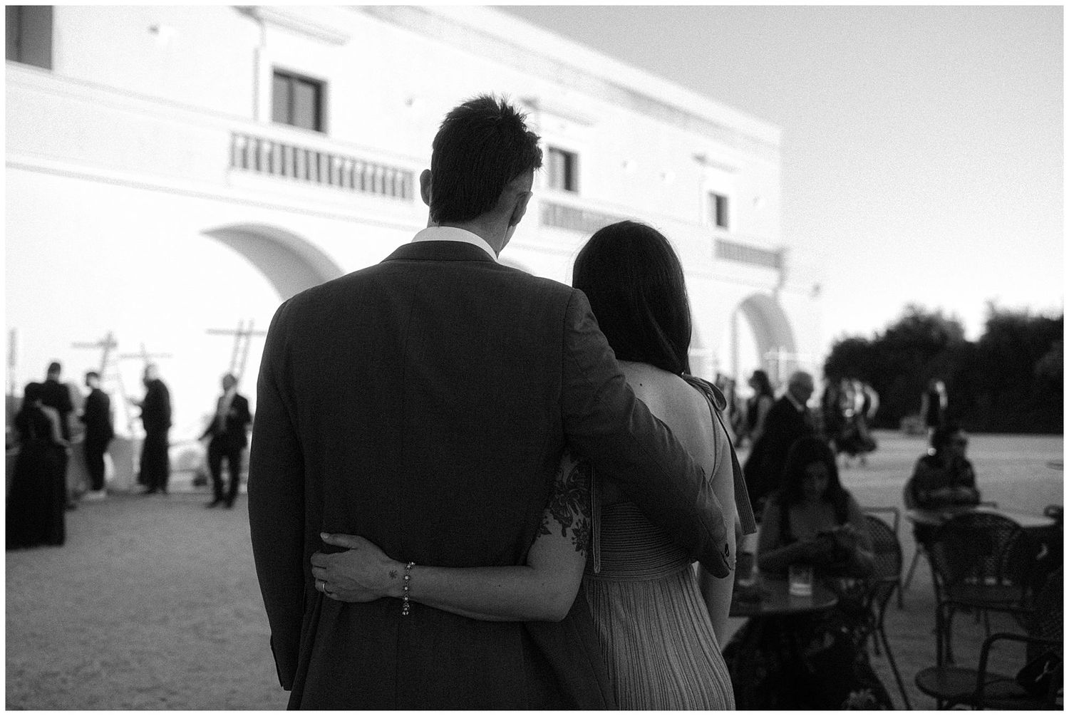 A couple embraces while watching guests at an outdoor wedding venue with a white building in the background.